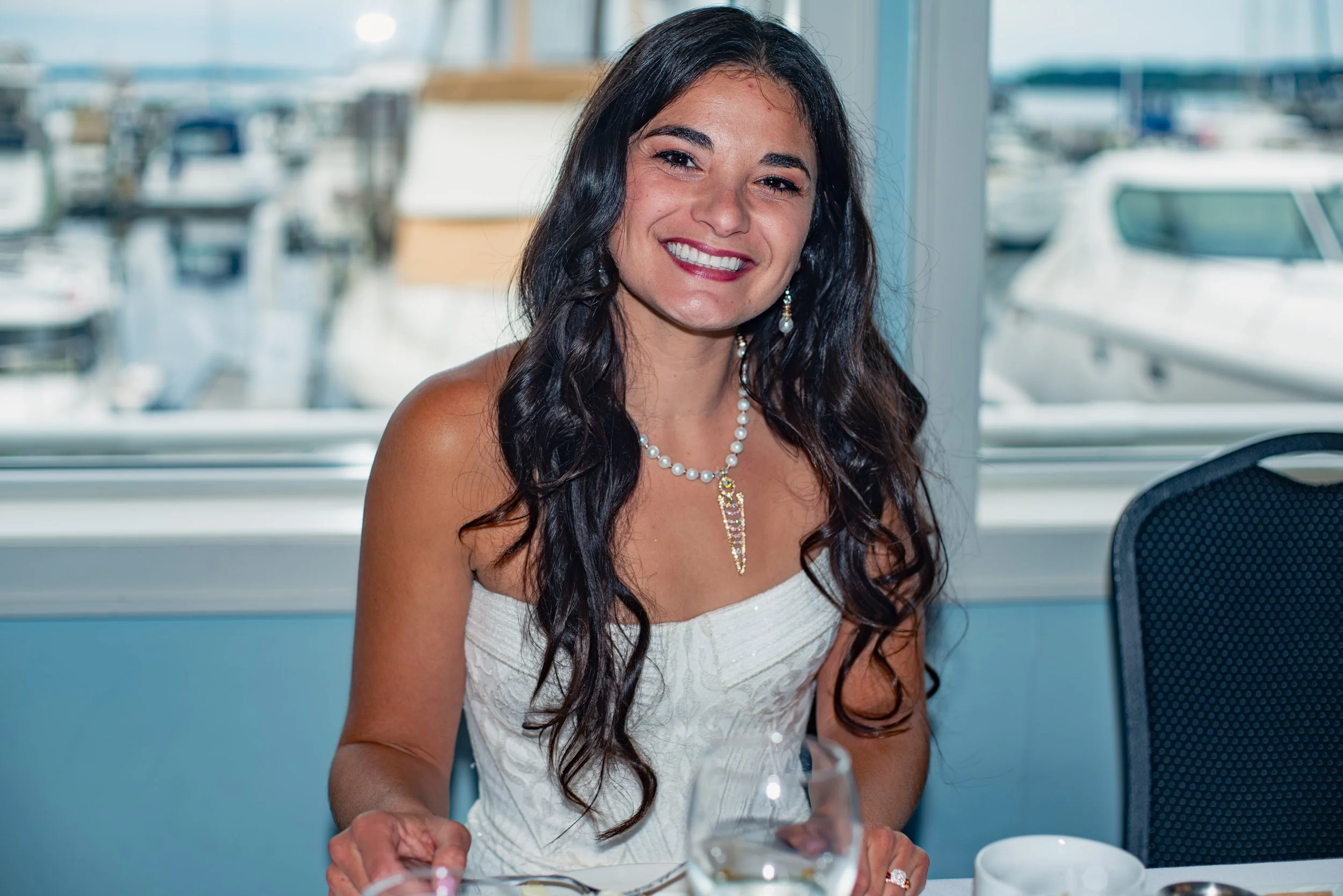 A woman with long dark wavy hair smiling at a table during a formal event, wearing a white dress and pearl jewelry, with a marina and boats visible through the window behind her.