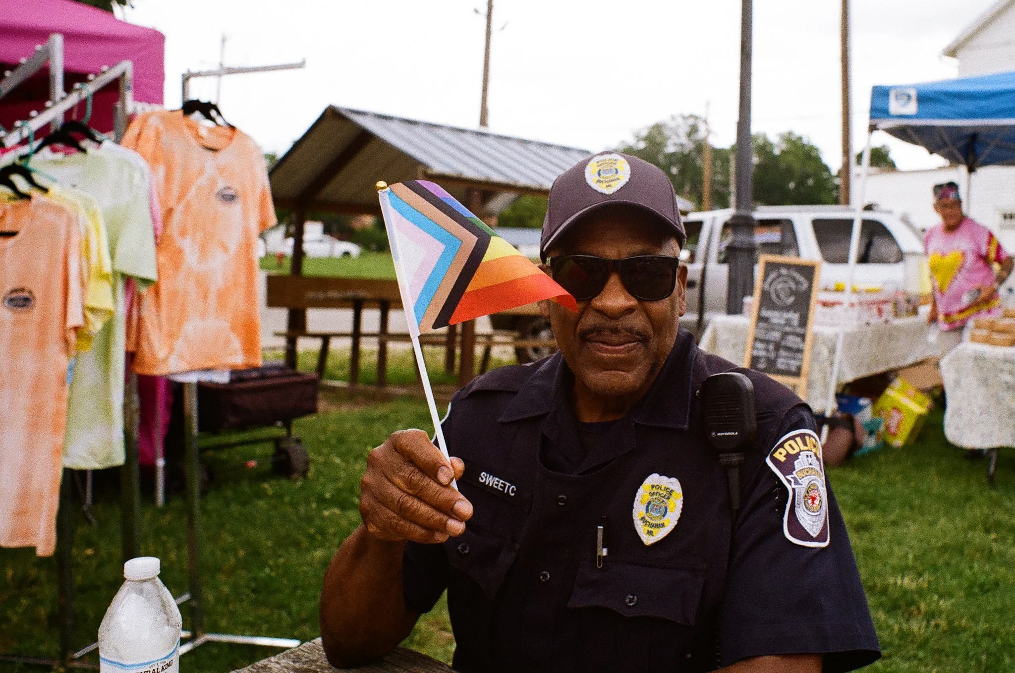 A police officer holding a colorful Pride flag at an outdoor event. The officer wears sunglasses, a police uniform with badge, and a hat. In the background, there are market stalls with clothing, a chalkboard sign, and a person in pink, with trees an