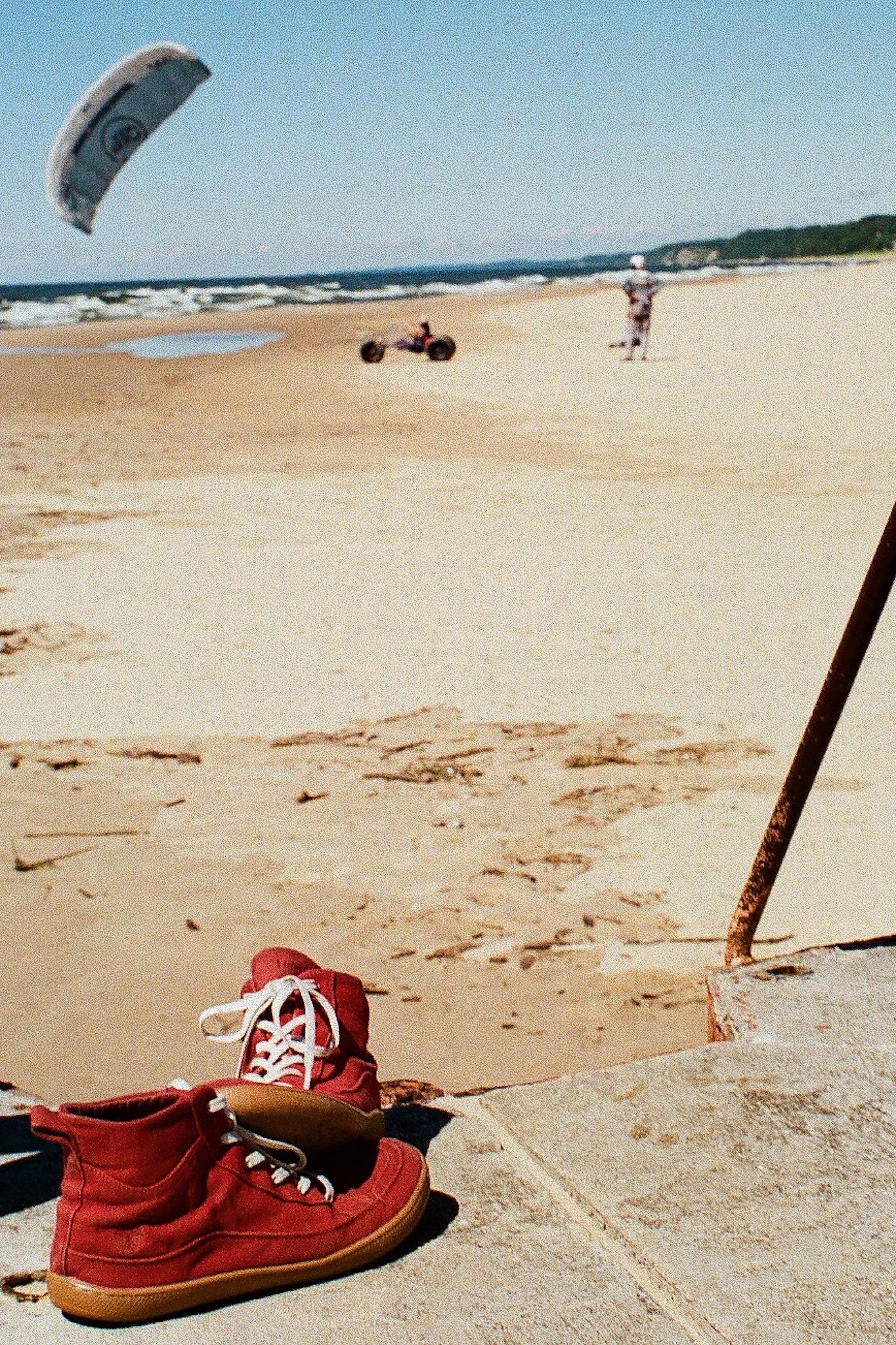 Red athletic shoes on sandy ground with kite surfing equipment and a person in the background near the ocean.