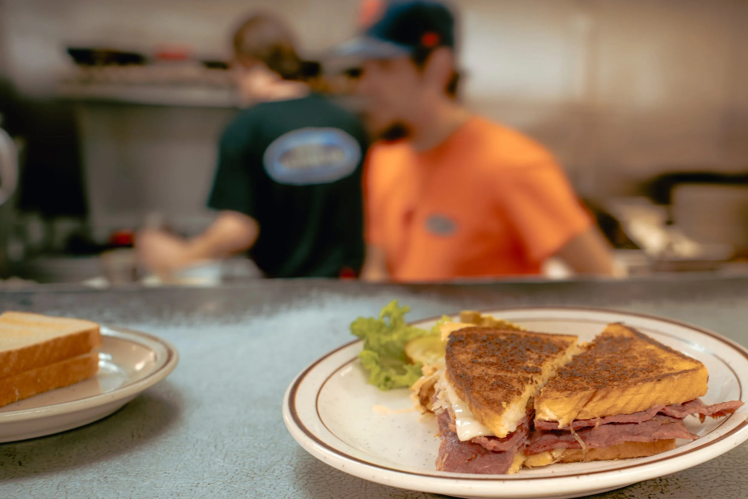 A close-up view of a plate with a club sandwich containing sliced turkey, ham, and cheese, garnished with lettuce and a pickle, in front of a blurred kitchen scene with a person wearing an orange shirt preparing food.