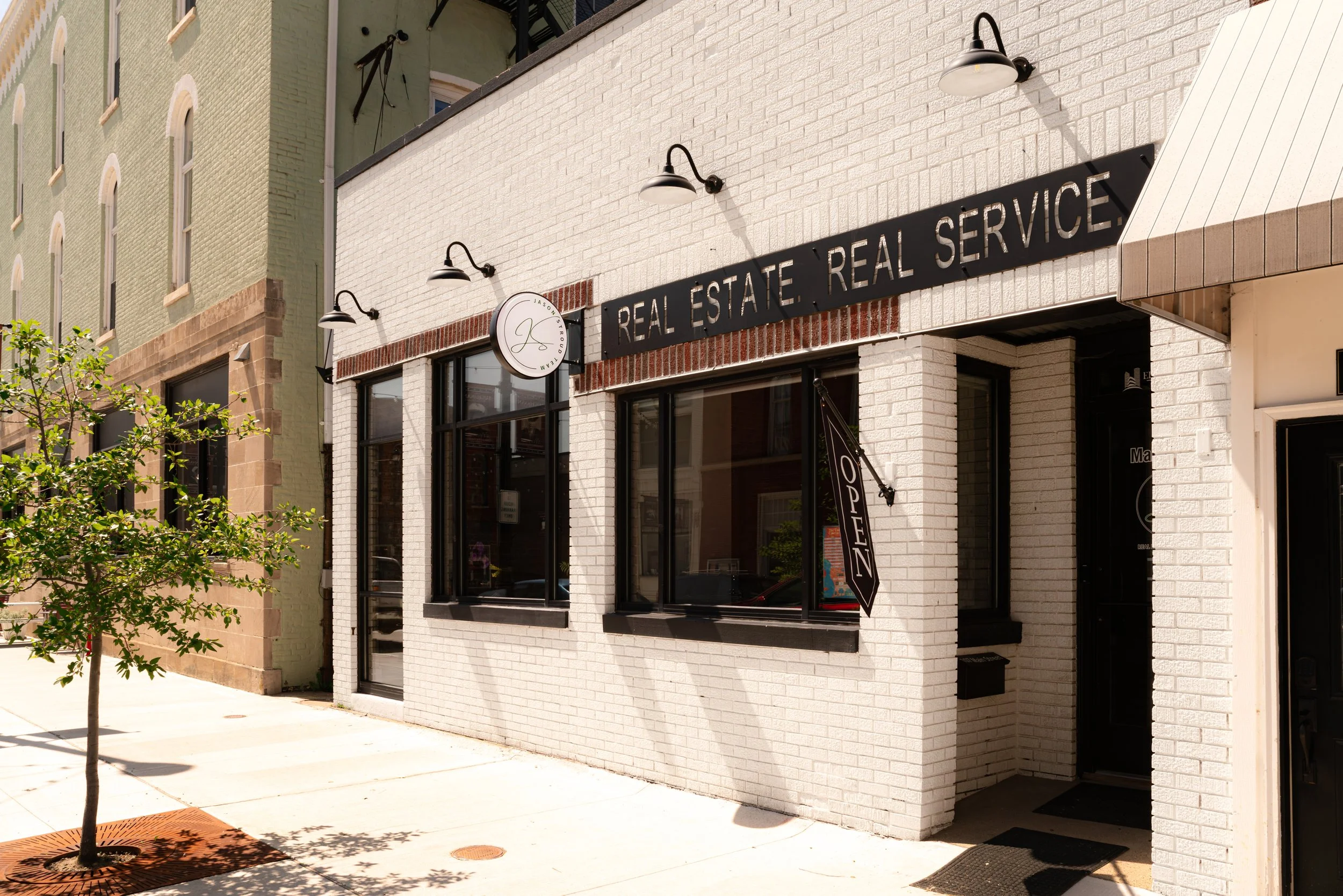 Exterior of a real estate office with a sign that reads 'Real Estate. Real Service.' The building has white brick walls with black-framed windows and a black hanging sign. There are two black outdoor light fixtures mounted on the wall above the sign.