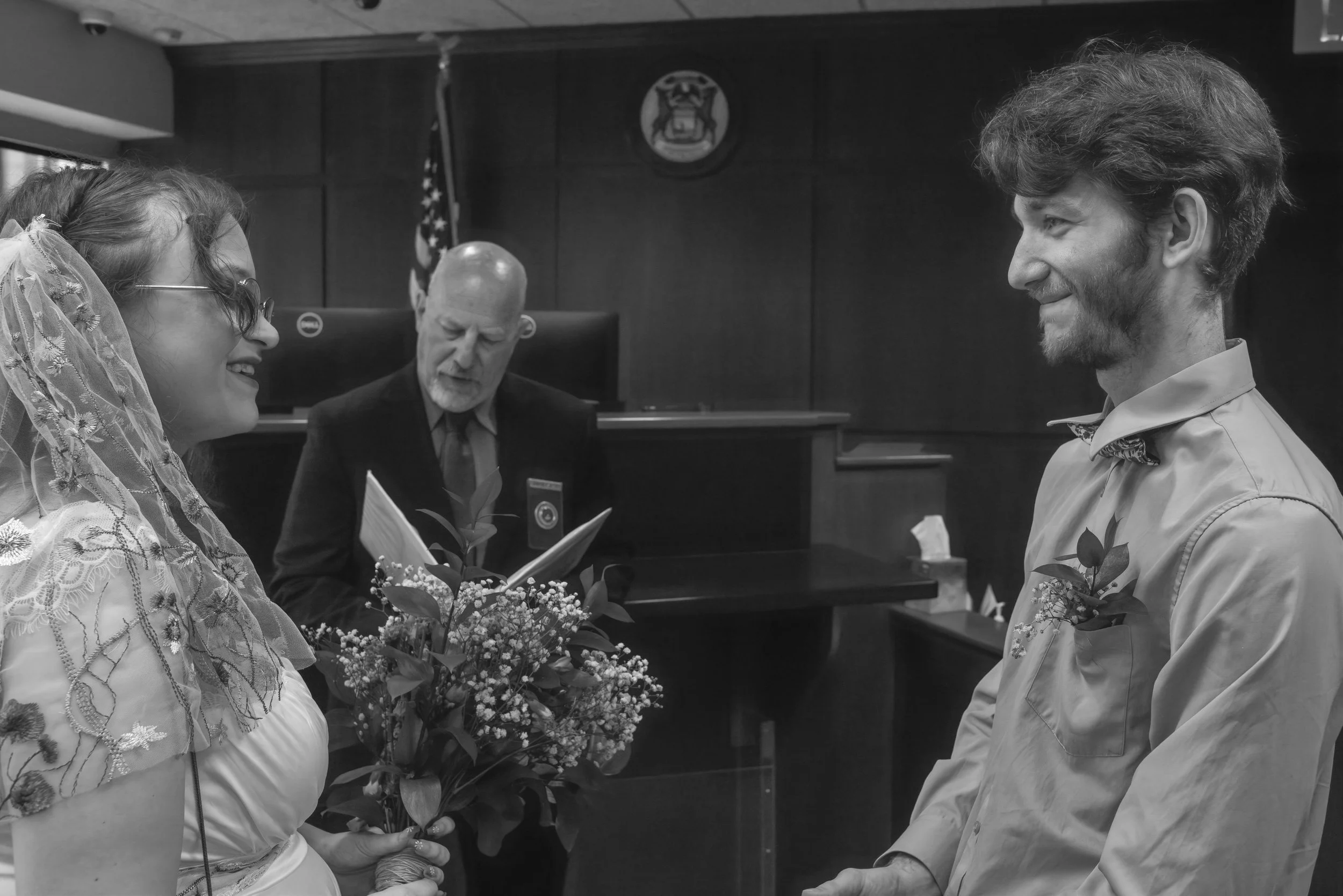 A black and white photo of a wedding ceremony in a courtroom. A bride and groom are facing each other, holding hands. The bride is wearing glasses and a veil, while the groom is wearing a boutonnière on his shirt. An officiant stands behind them, rea