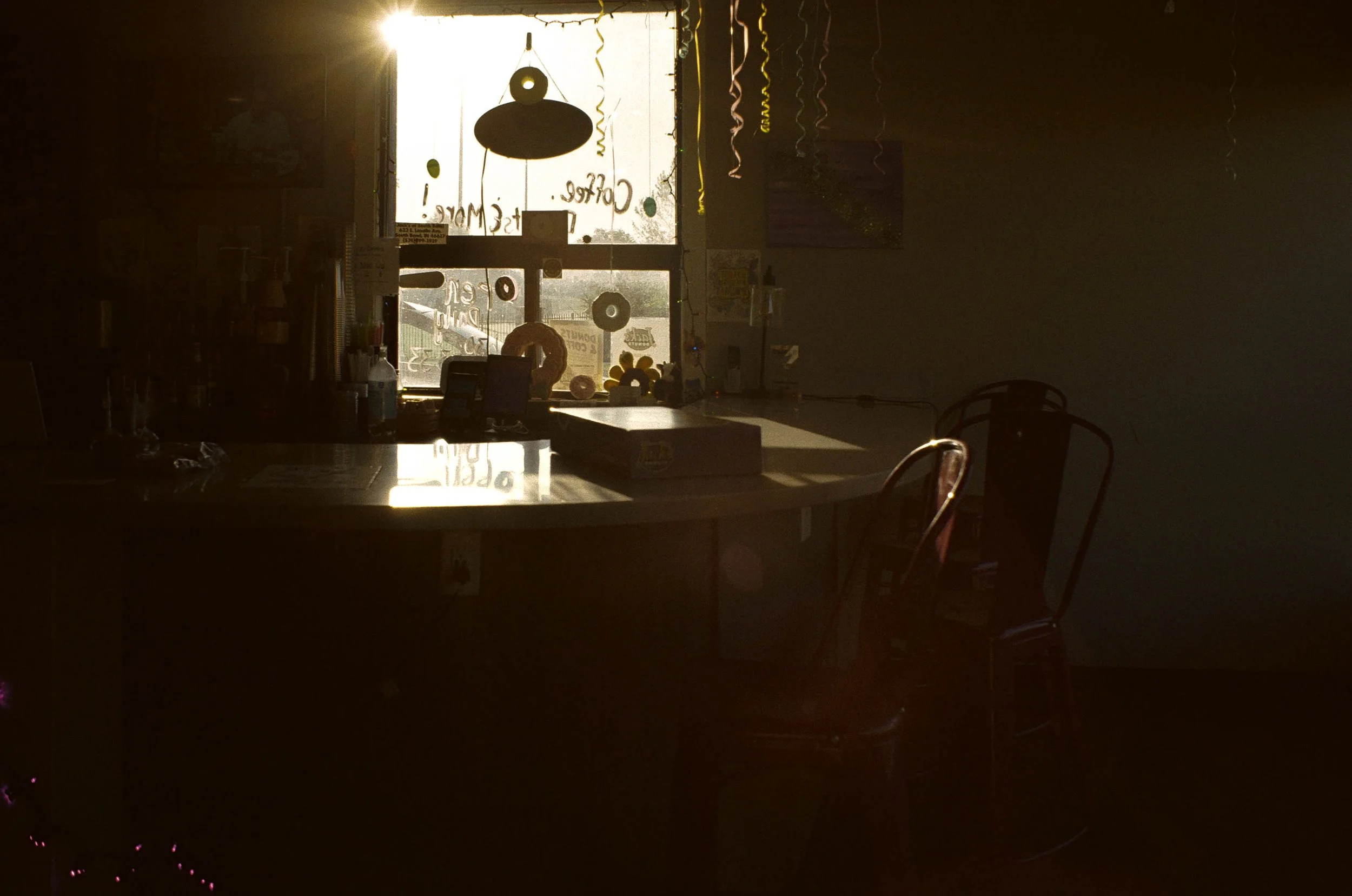 A dimly lit room with a large window in the background. The window has signs and decorations, including streamers and a donut-shaped object. A table in the foreground displays various items, and chairs are partially visible.