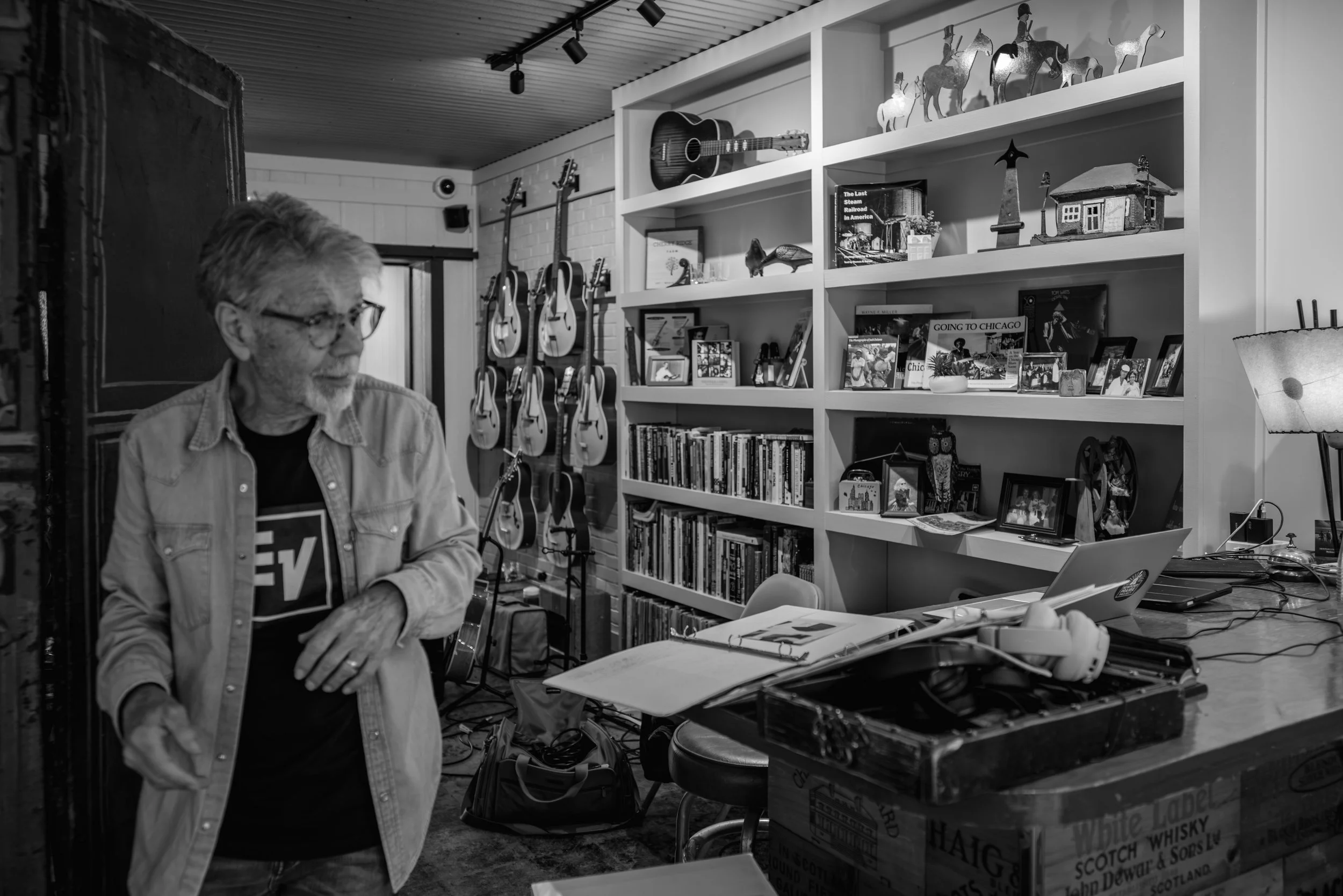 A man with glasses and a beard, wearing a denim jacket, stands at a cluttered table in a room filled with guitars on the wall and bookshelves. The room has a corrugated metal ceiling and various decorative items.