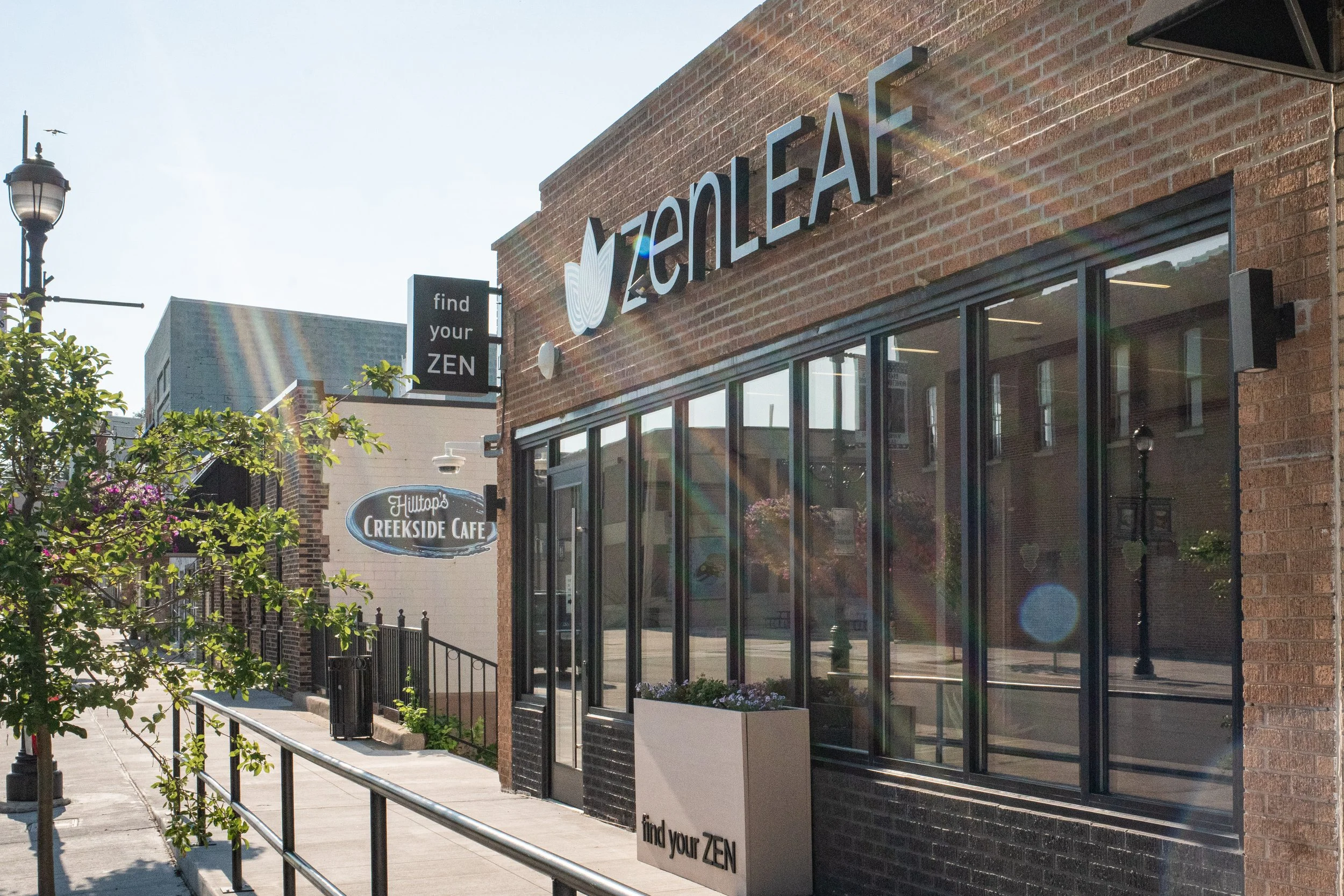 Street view of a brick building with a sign reading 'Zenergy', window display, and a nearby sign that says 'find your ZEN'. There are trees, a street lamp, and other buildings in the background.
