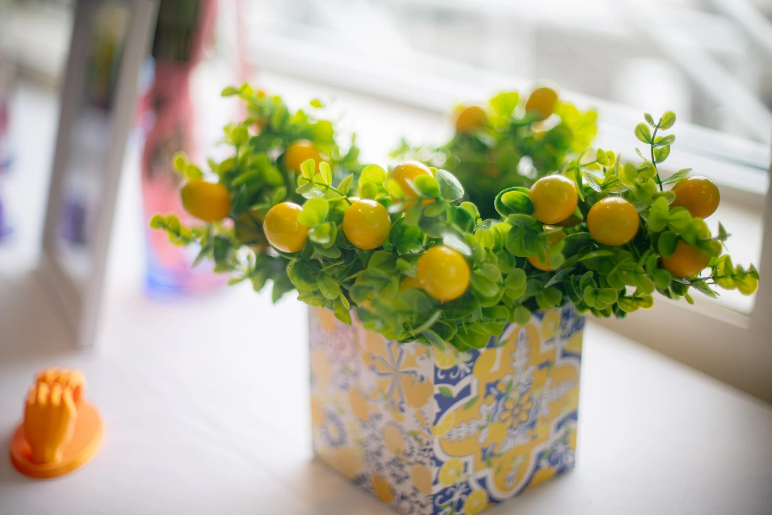 A potted plant with small yellow fruits and green leaves on a windowsill, with blurred colorful objects in the background.