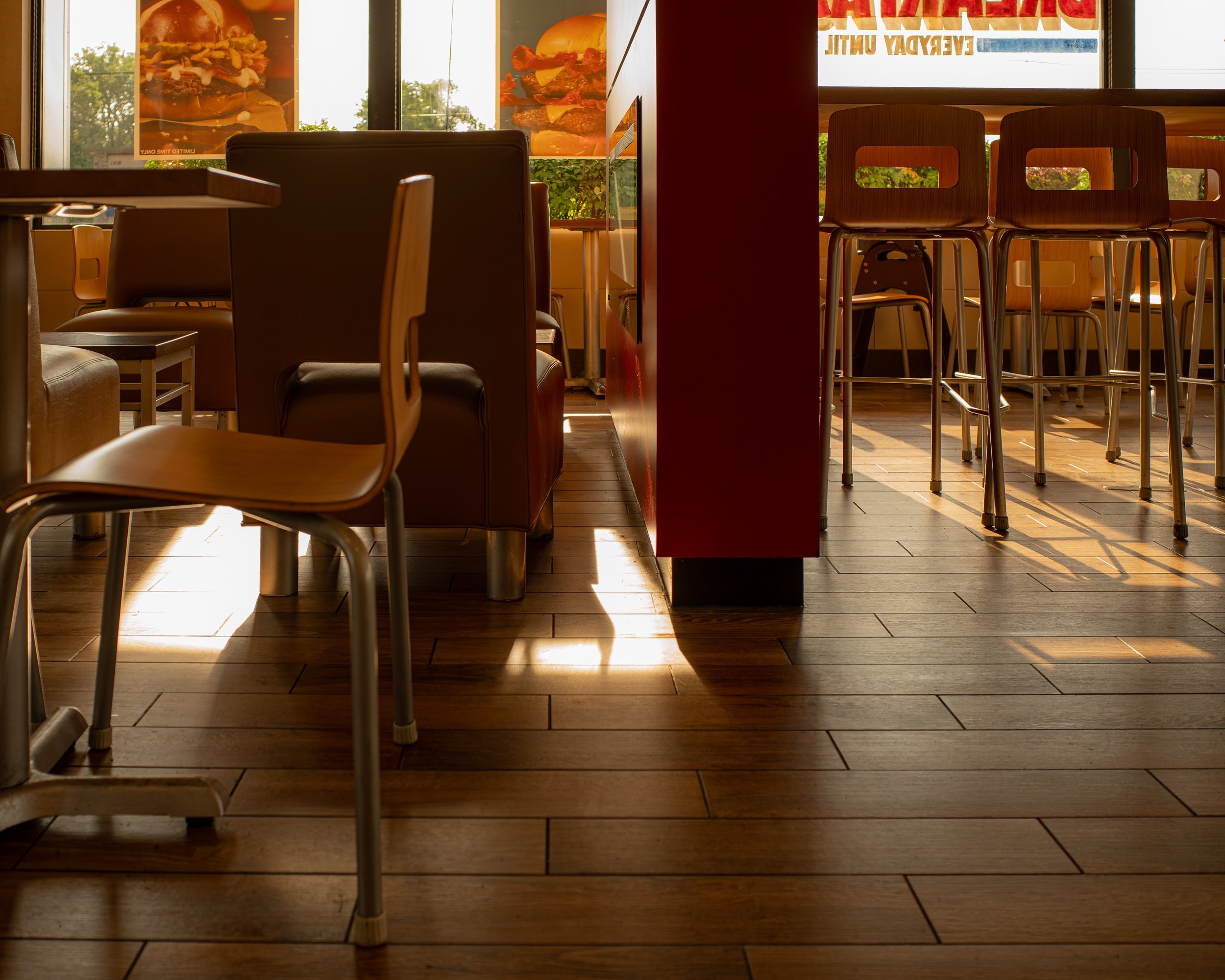 Empty restaurant with wooden chairs and tables, sunlight streaming through large windows with advertising posters of hamburgers.