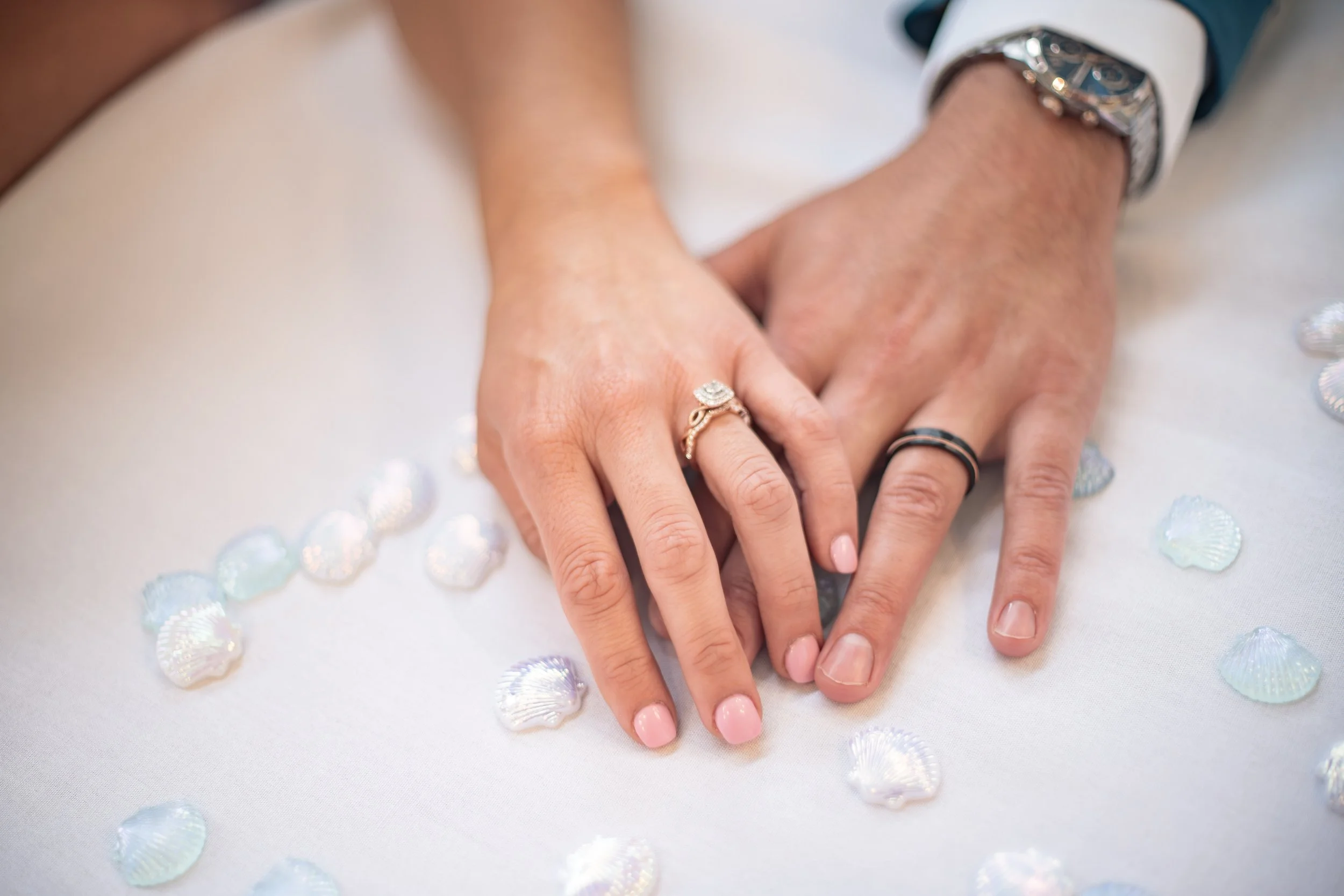 Close-up of hands, one with a silver watch and rings, on a white surface with scattered seashells.