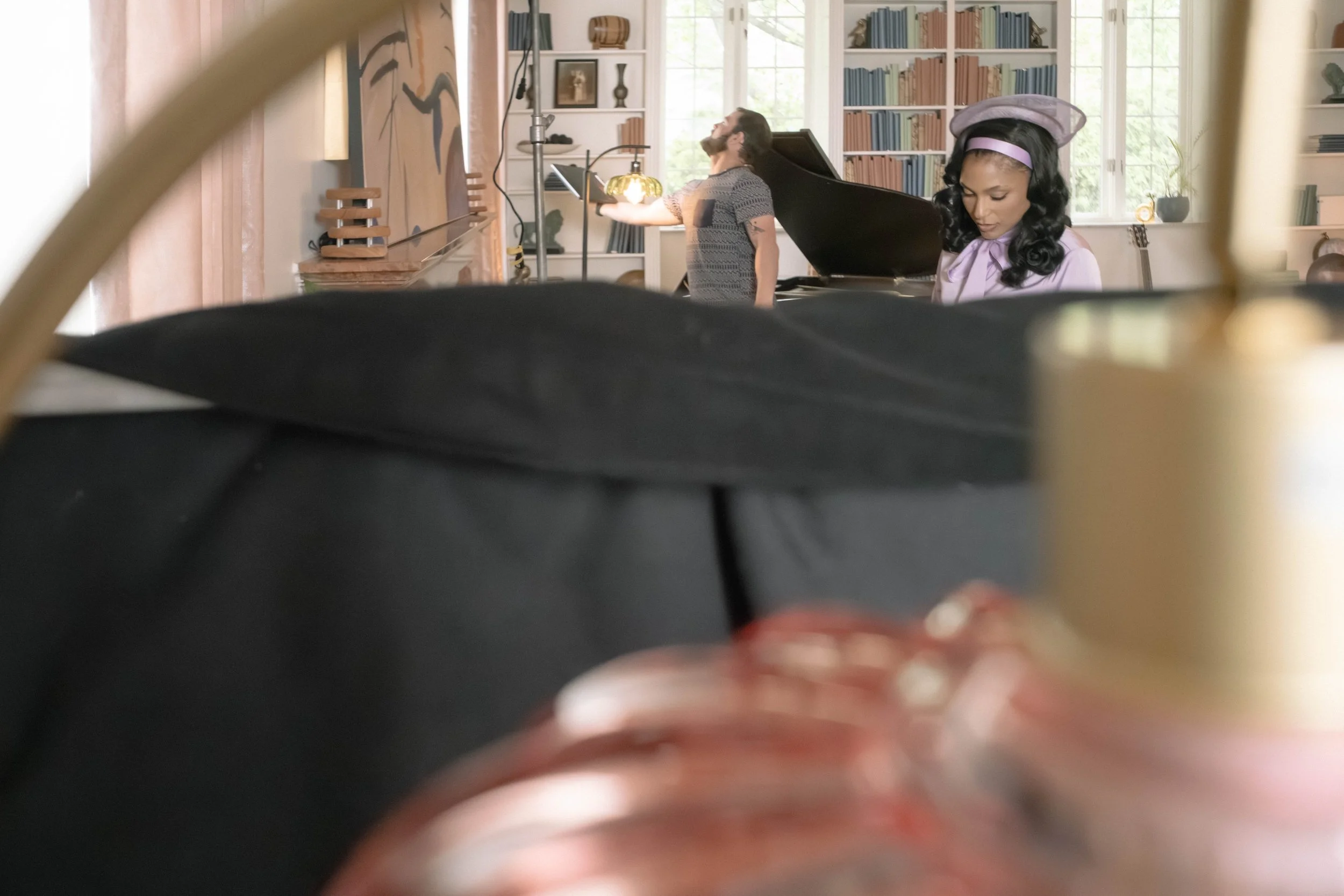 People in a bright room with bookshelves, a man singing or speaking passionately, and a woman with vintage attire and a hair net, possibly preparing for a performance.