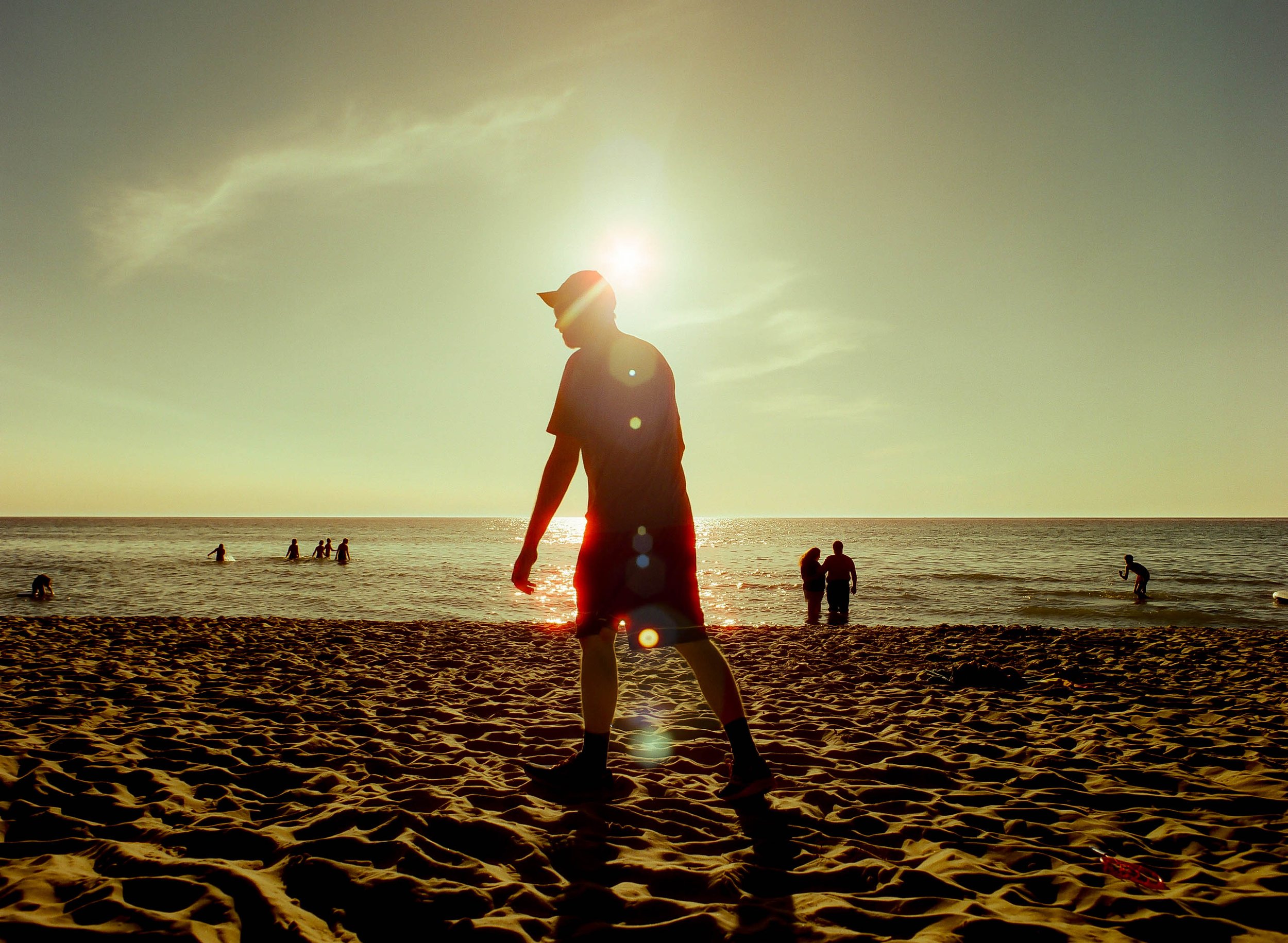 A person wearing a hat and shorts walking on a sandy beach during sunset, with several people in the water and on the beach, and a cloudy sky in the background.
