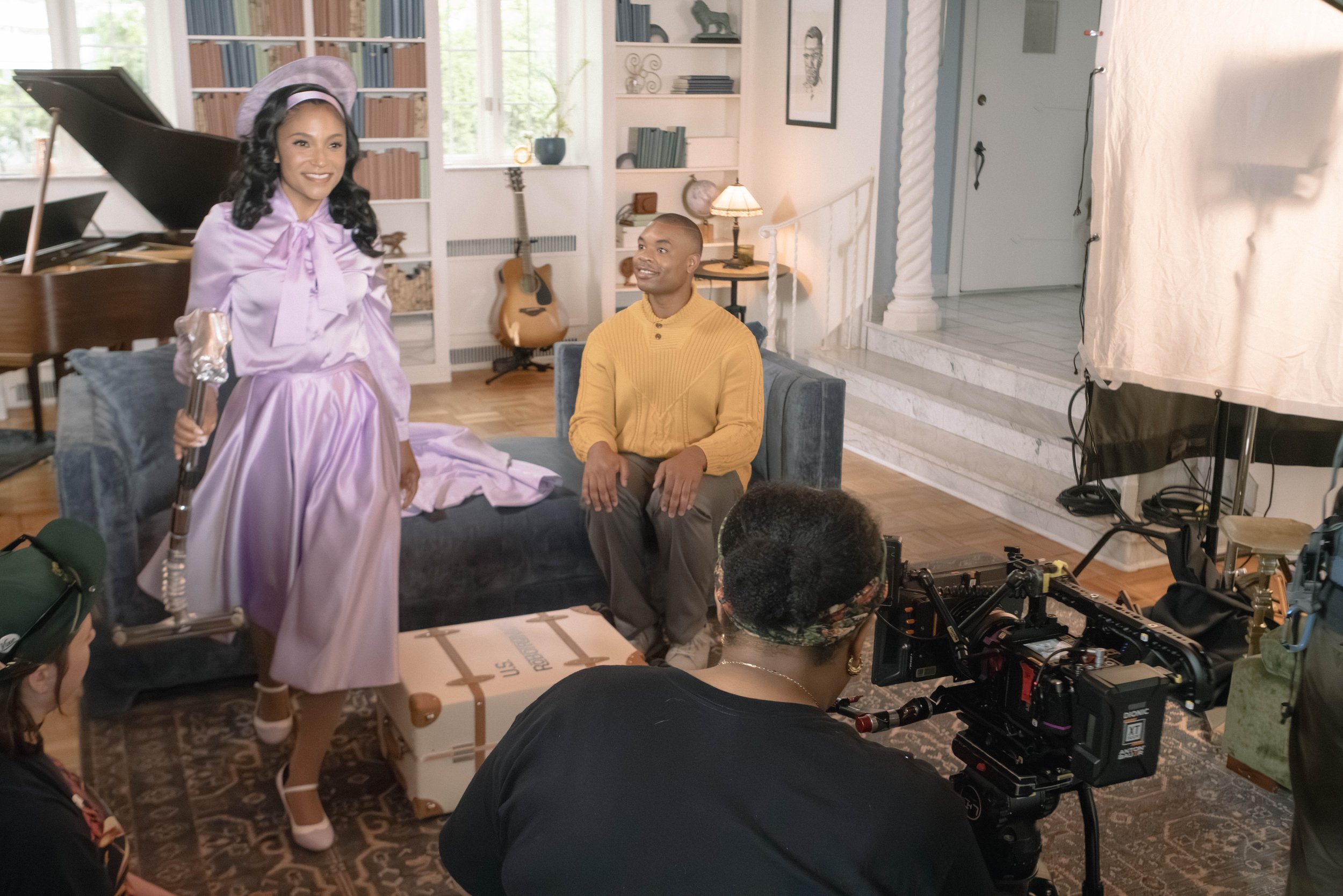 A woman dressed in a lavender vintage style dress and hat holds a microphone while standing in a room with a man seated on a couch and crew members operating camera equipment. The room has a piano, bookshelves, and large windows.