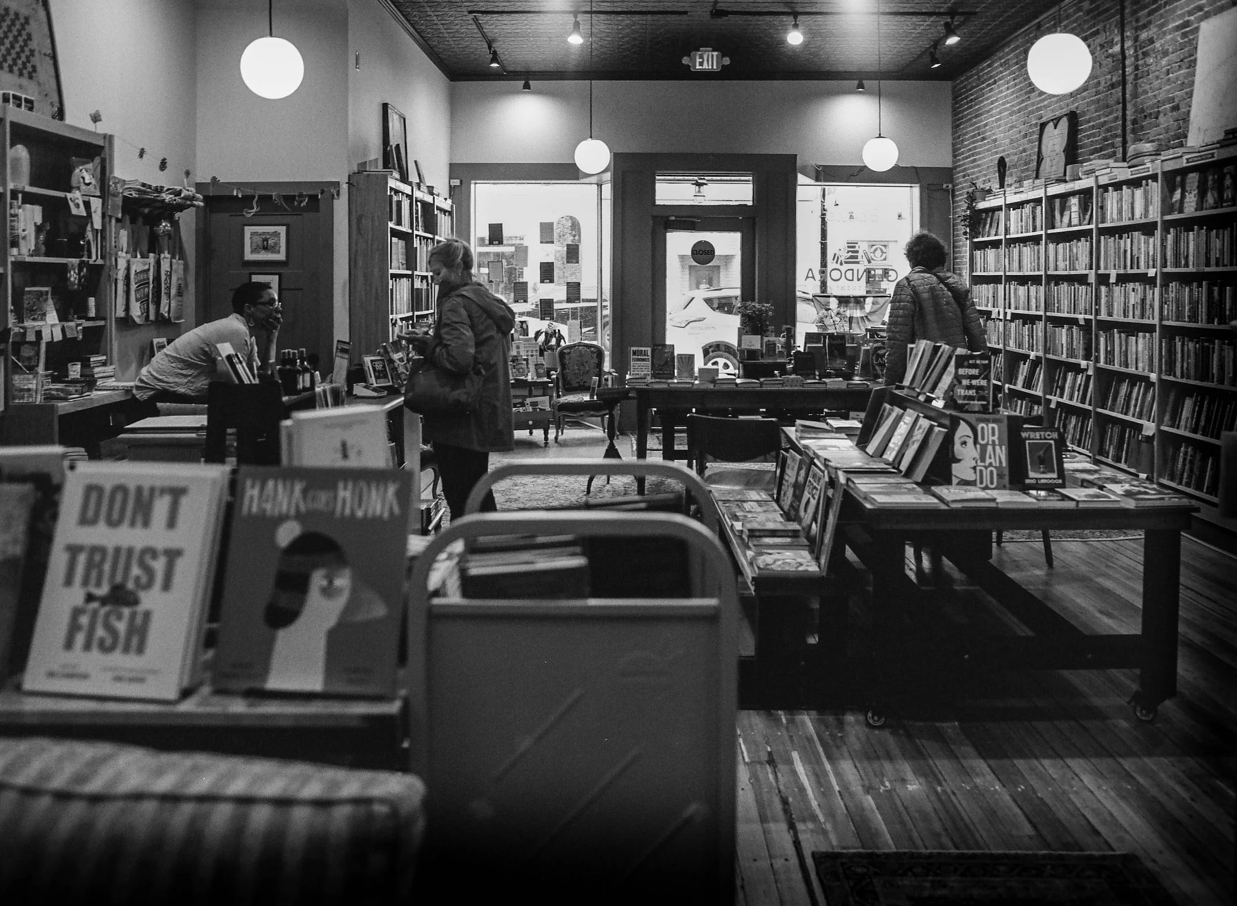 Black and white photo of the interior of a bookstore with shelves filled with books, two women browsing, and a man sitting at a table, with large windows and an 'EXIT' sign.