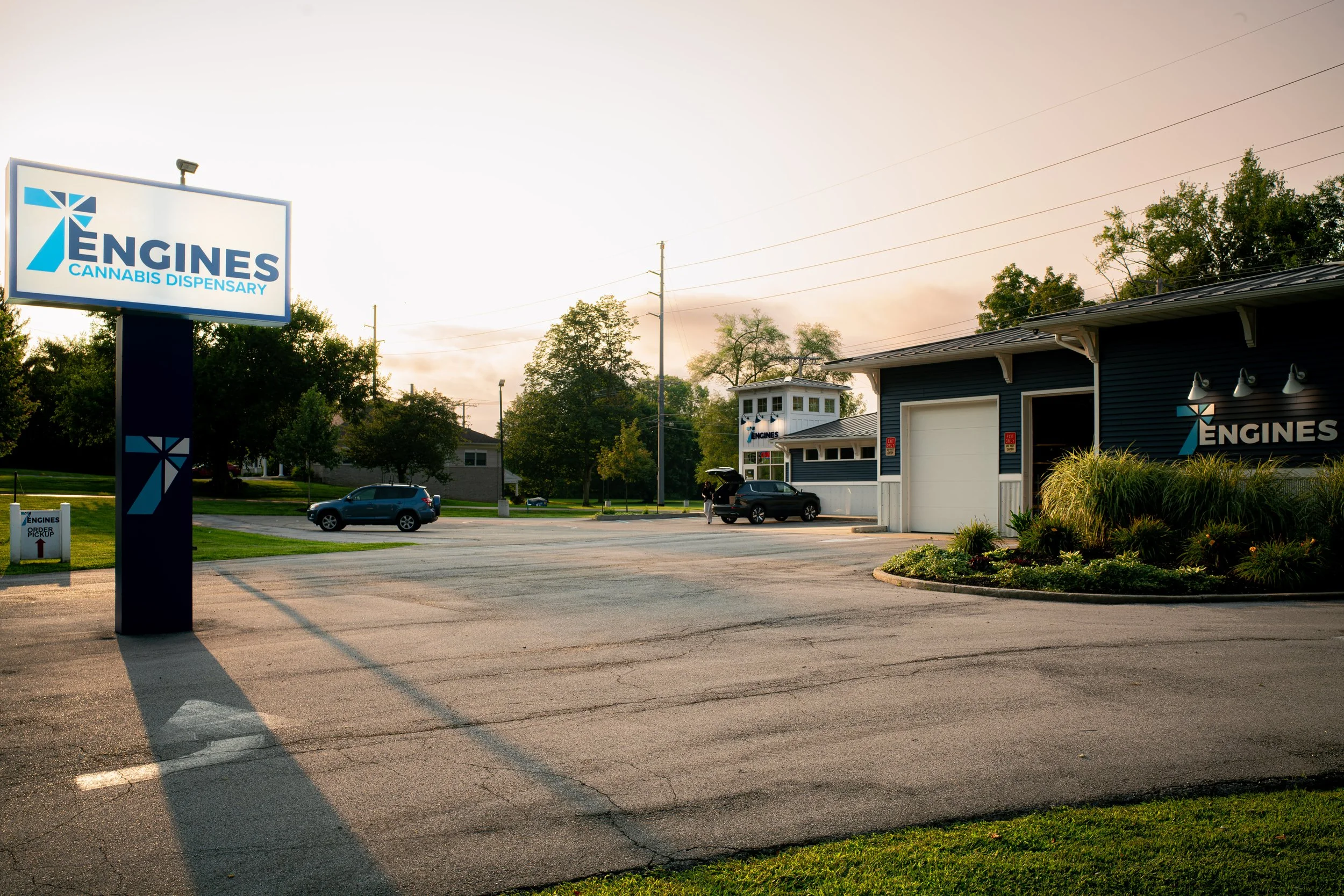 An outdoor view of a cannabis dispensary named 'Engines', with a parking lot, a blue building, and a lit signboard displaying the dispensary's name and logo at sunset.