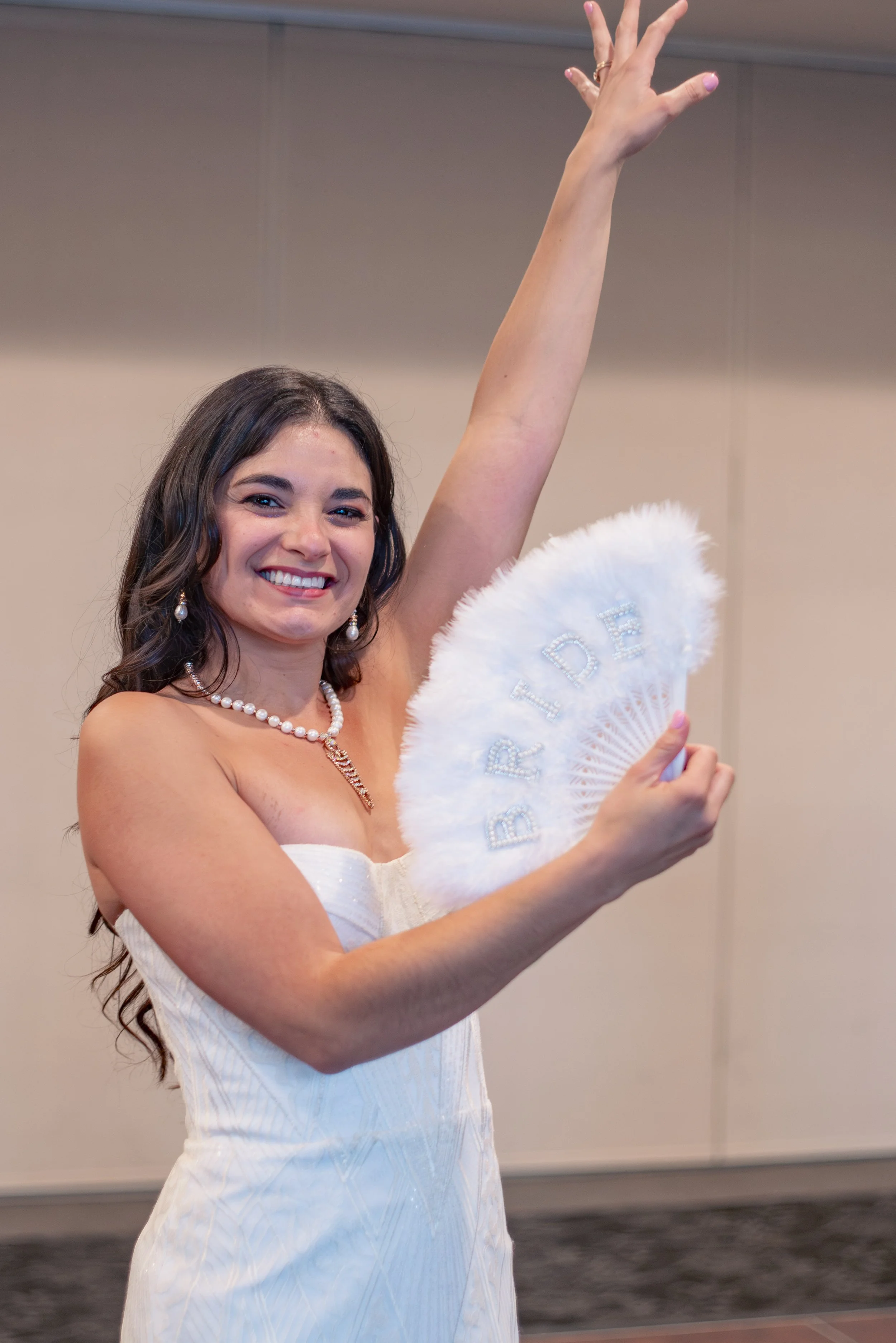 A woman with long dark hair and a bright smile wearing a white dress, pearl necklace, and pearl earrings, raising her right arm and holding a white fan with the word 'BRIDE' embroidered on it.