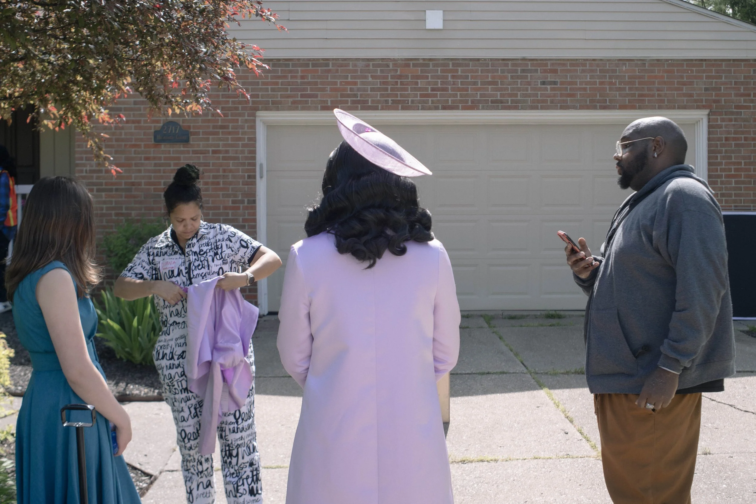 People gather outside on a sunny day in front of a brick house with a garage. There are four women and one man visible. One woman is dressed in a purple outfit with a matching purple hat, and others wear casual dresses and clothing. The man has a bea
