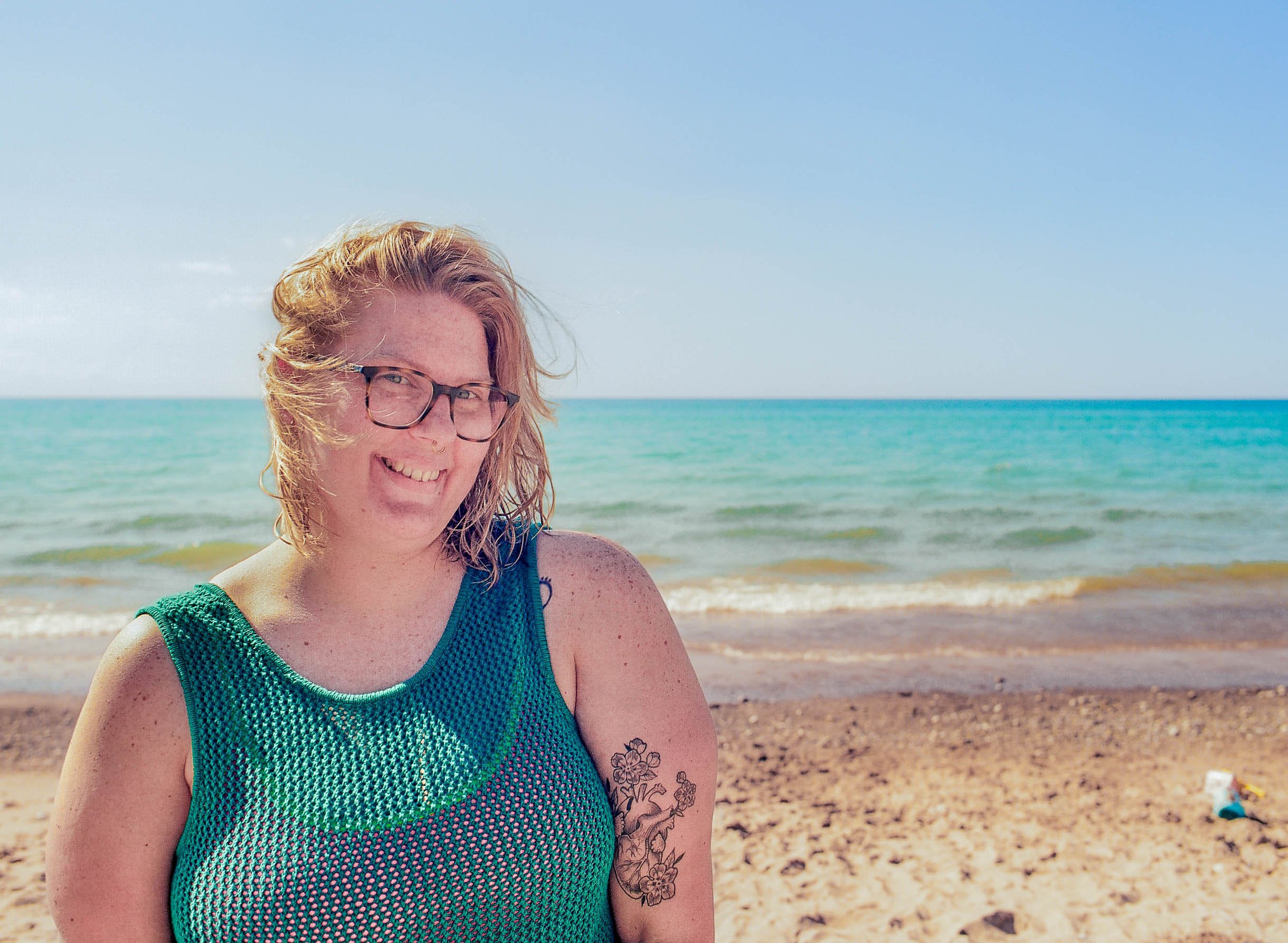 A woman with blonde hair, glasses, and tattoos on her arm, smiling at the beach with the ocean and sky in the background.