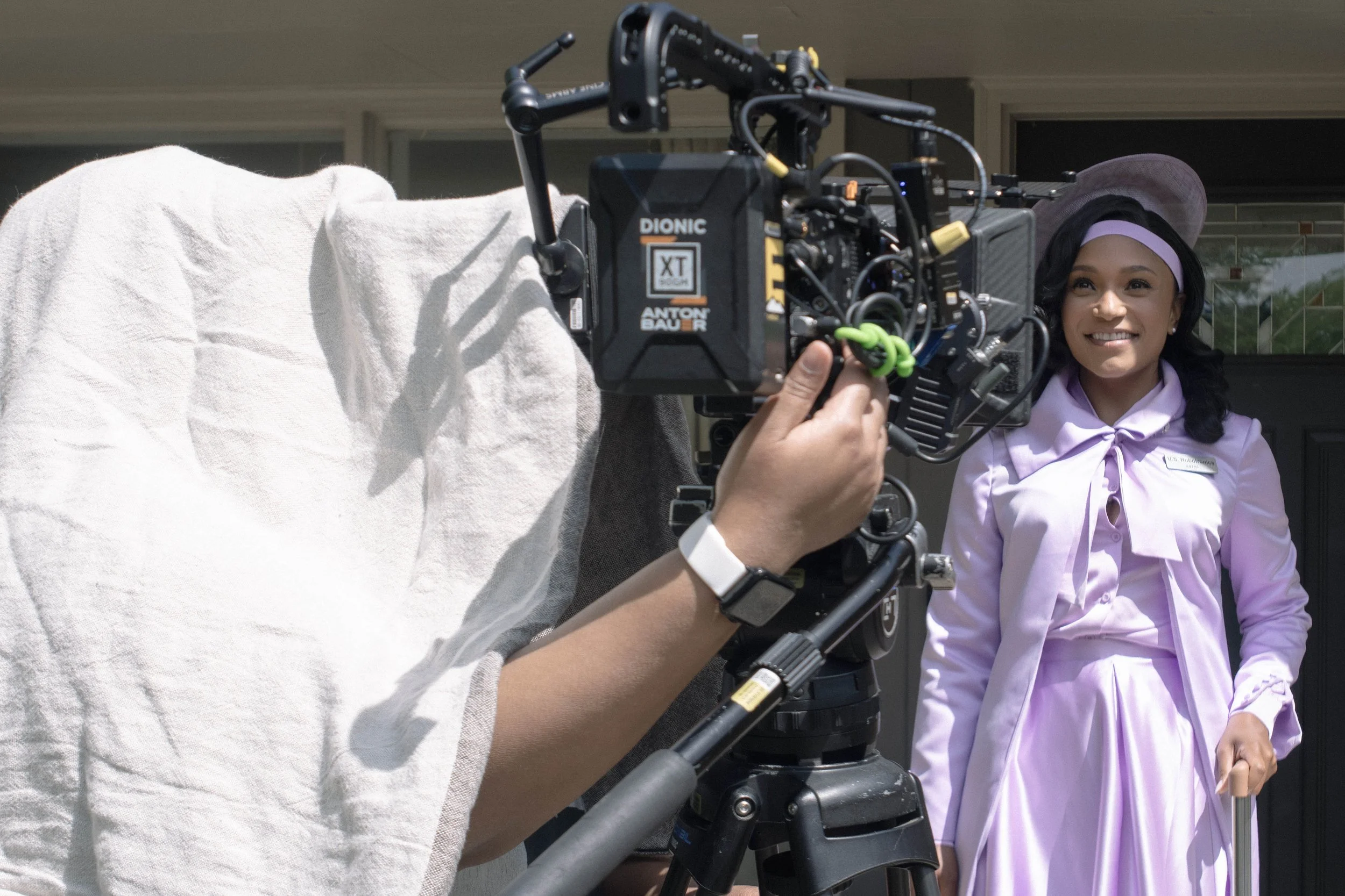 A woman with long dark hair, wearing a purple hat and light purple blouse, smiling while standing outside. Camera equipment is focused on her, capturing her portrait against a building background.