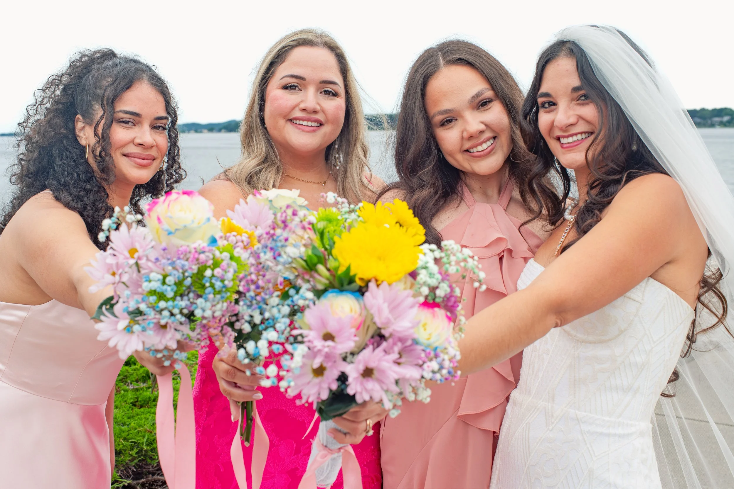 Four women smiling at the camera, holding a bouquet of colorful flowers, with a body of water and a distant shoreline in the background. One woman is wearing a wedding veil.