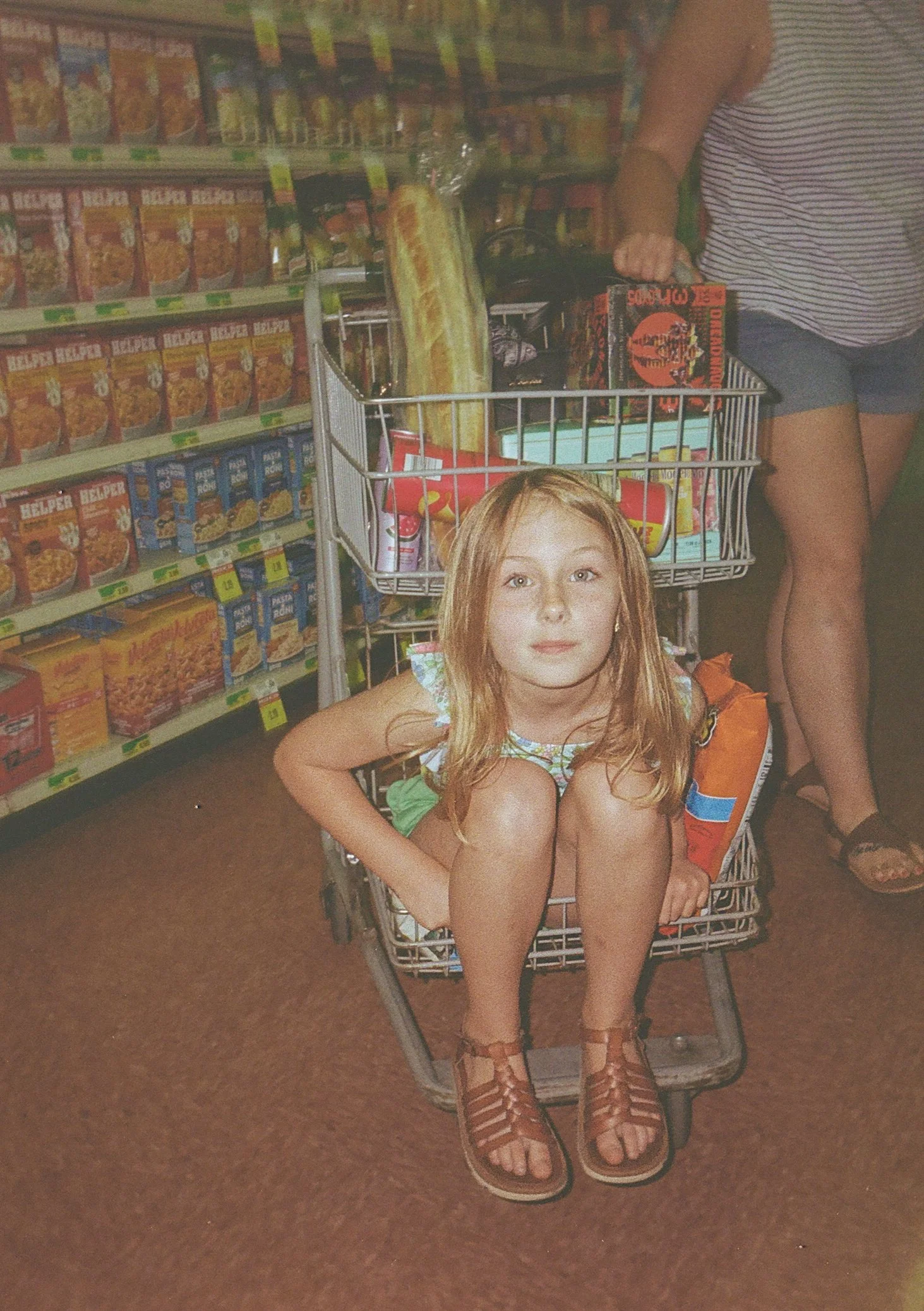 A young girl with red hair sitting inside a shopping cart in a grocery store aisle, surrounded by shelves of food products.