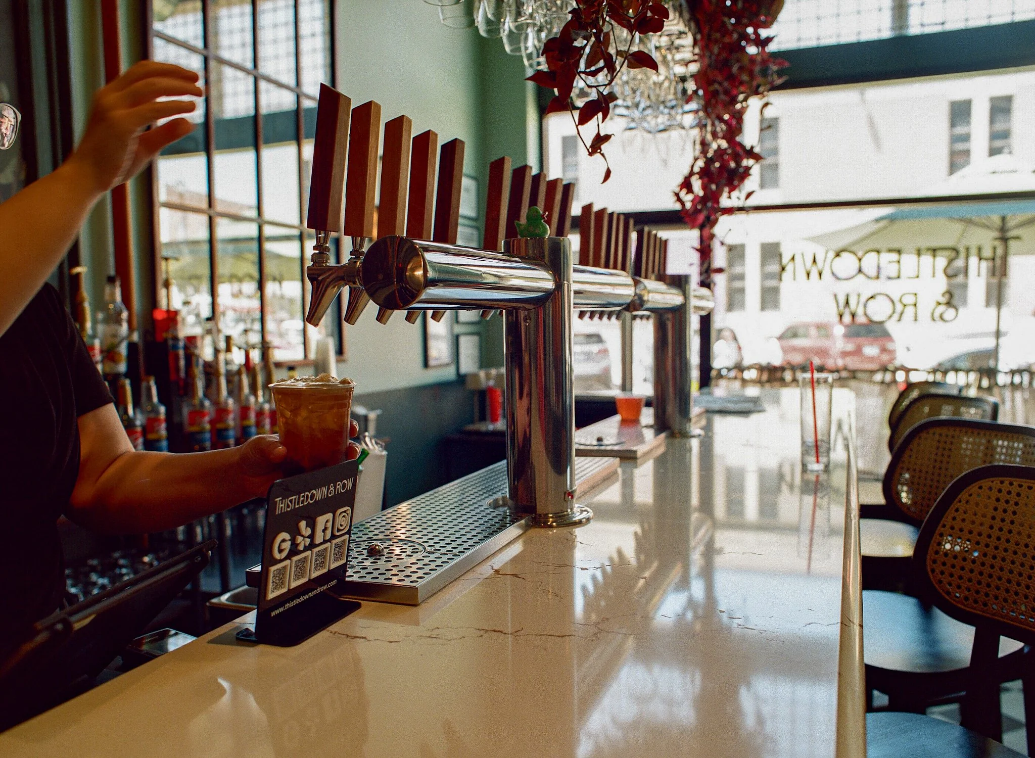 Person hands holding a cup of coffee or tea at a counter in a cafe with a beer tap, colorful decorations, and large windows in the background.
