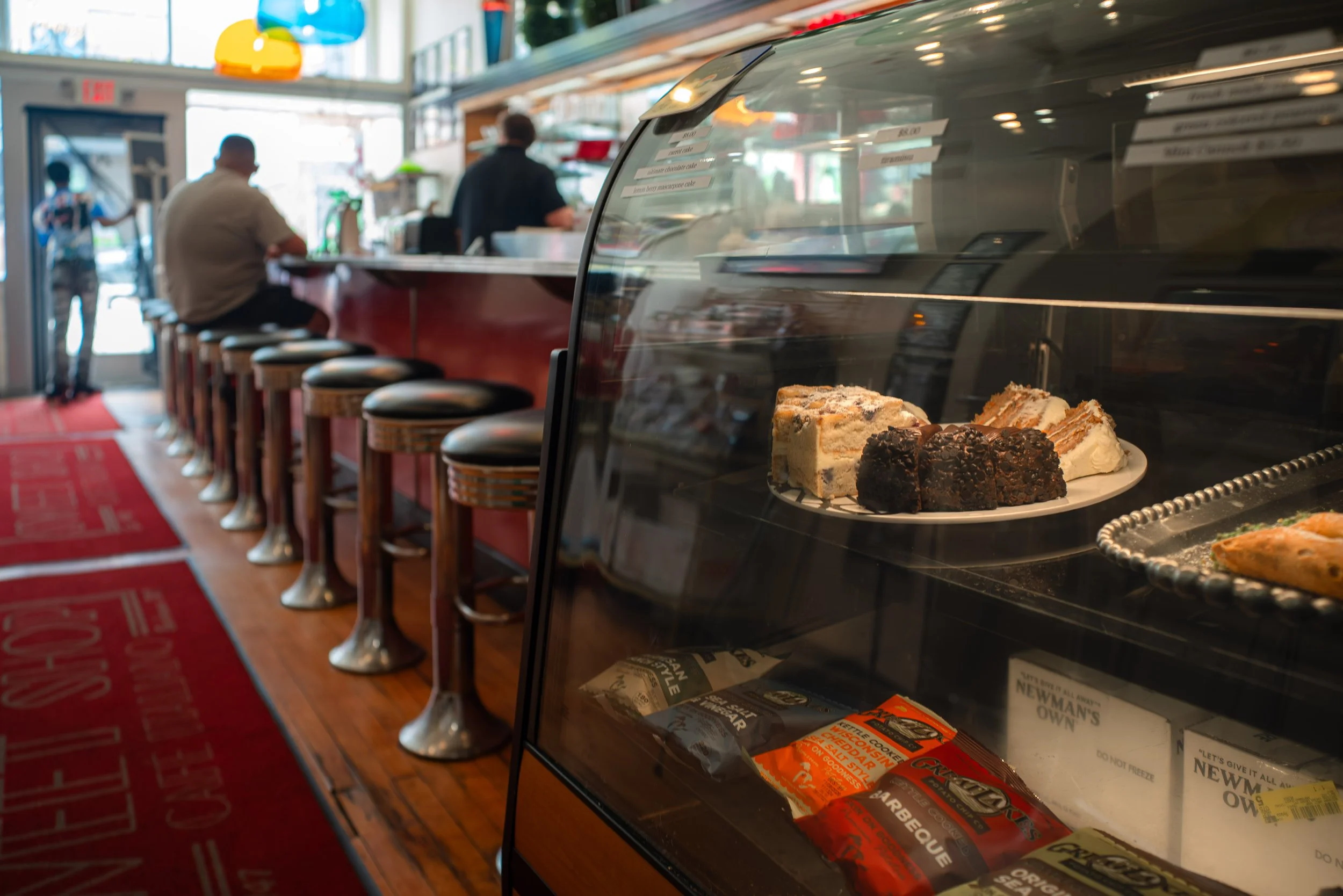 A pastry display case with slices of cake and baked goods inside, and a sandwich or pizza on a plate below. In the background, there are several customers ordering at a counter in a casual bakery or cafe.