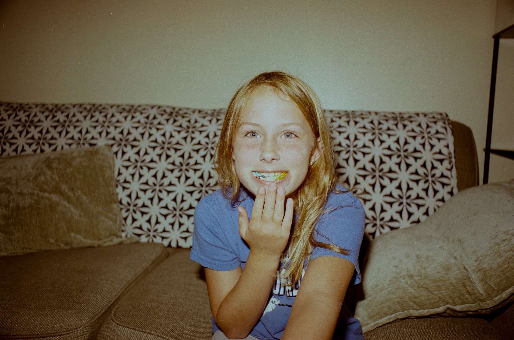A young girl with red hair and freckles smiling on a brown sofa in a living room.