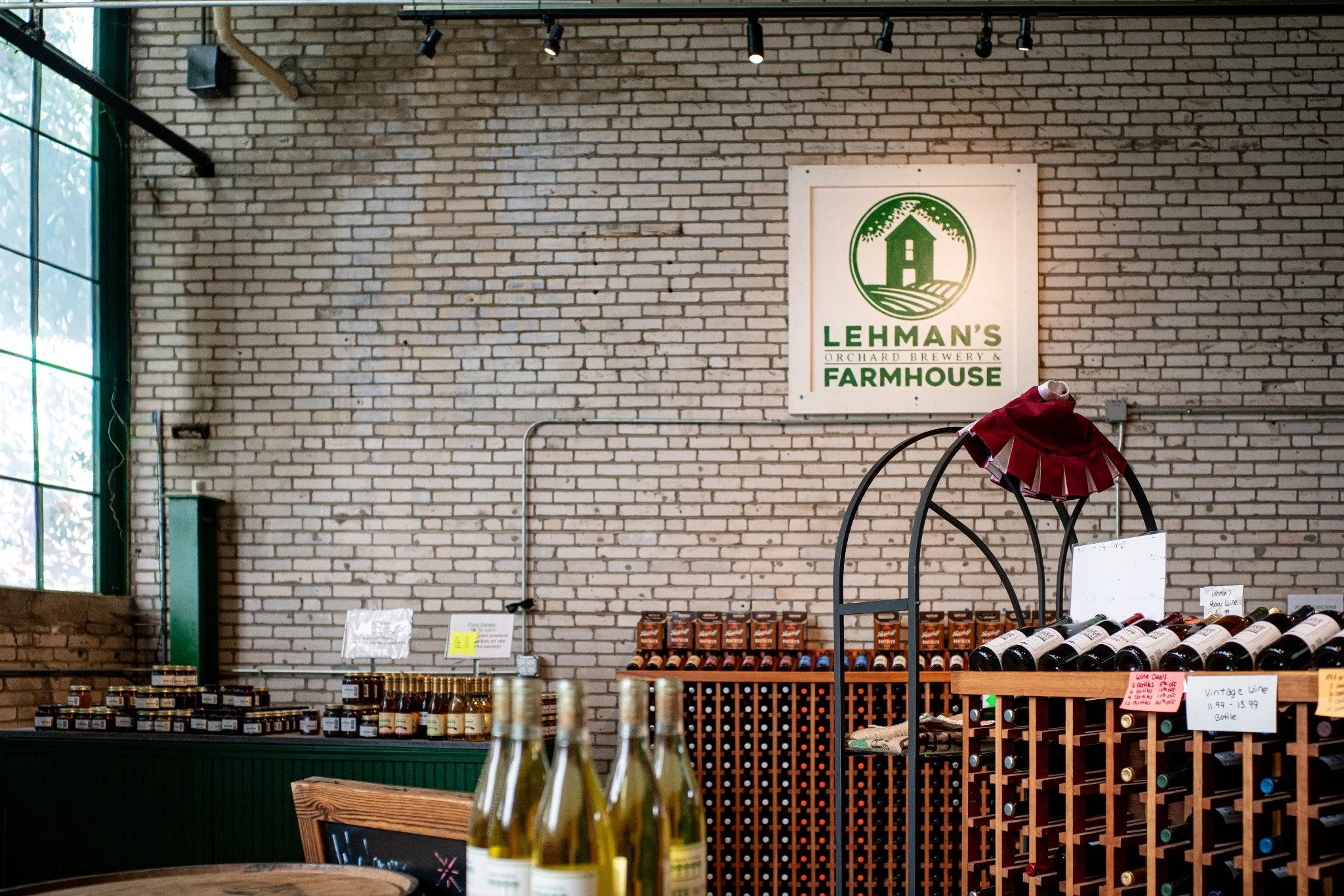 Interior view of Lehman's Orchard Brewery & Farmhouse with wine bottles on display, a brick wall, a Lehman's sign, and some bottles in the foreground.