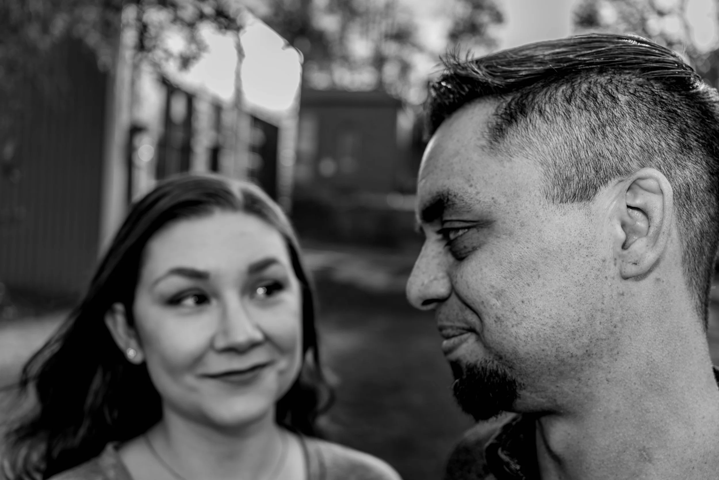 Close-up black and white photo of a man and woman looking at each other outdoors, smiling, with blurred trees and sky in the background.