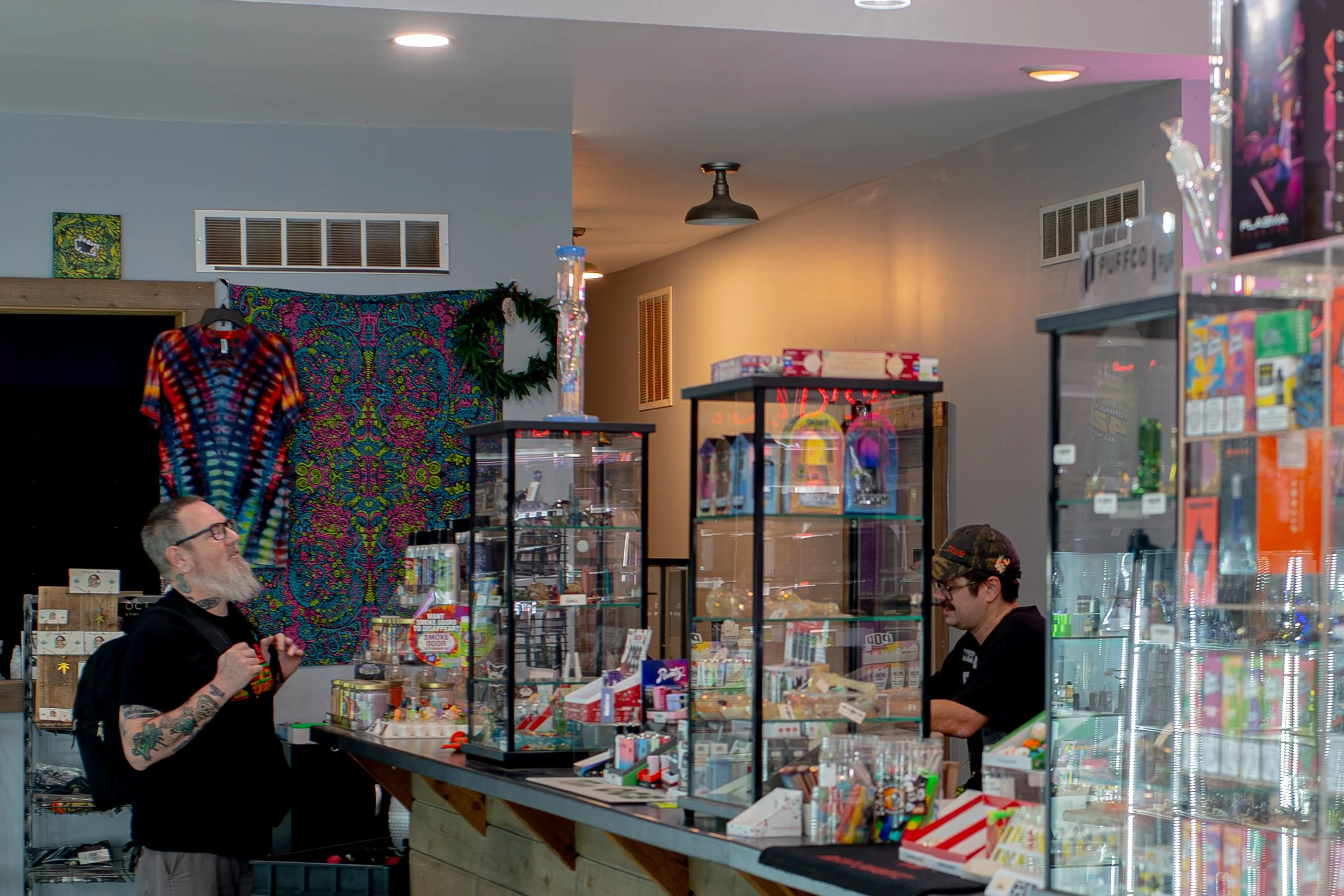 Inside a retail store with glass display cases filled with miscellaneous items, two shoppers, one with a tattooed arm wearing glasses and another with a cap, are browsing.