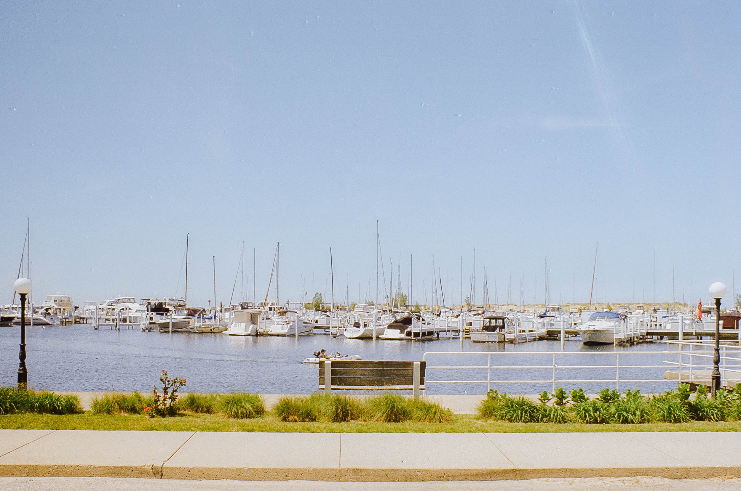 A marina with boats docked at the pier under a clear sky, with lamp posts and a landscaped area in the foreground.