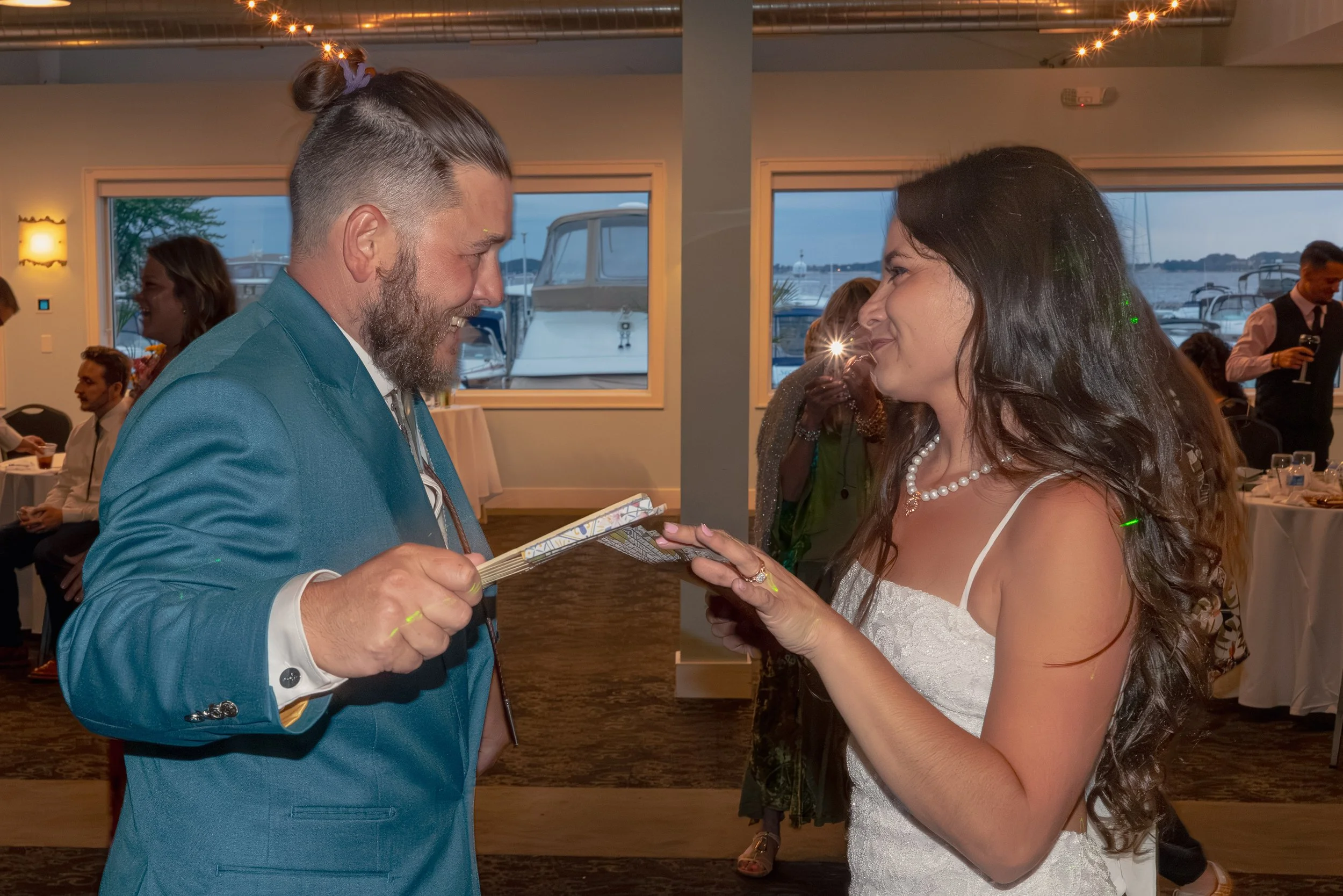 A man in a blue suit and a woman in a white dress exchange wedding vows at a celebration by the water, with boats visible through windows in the background.