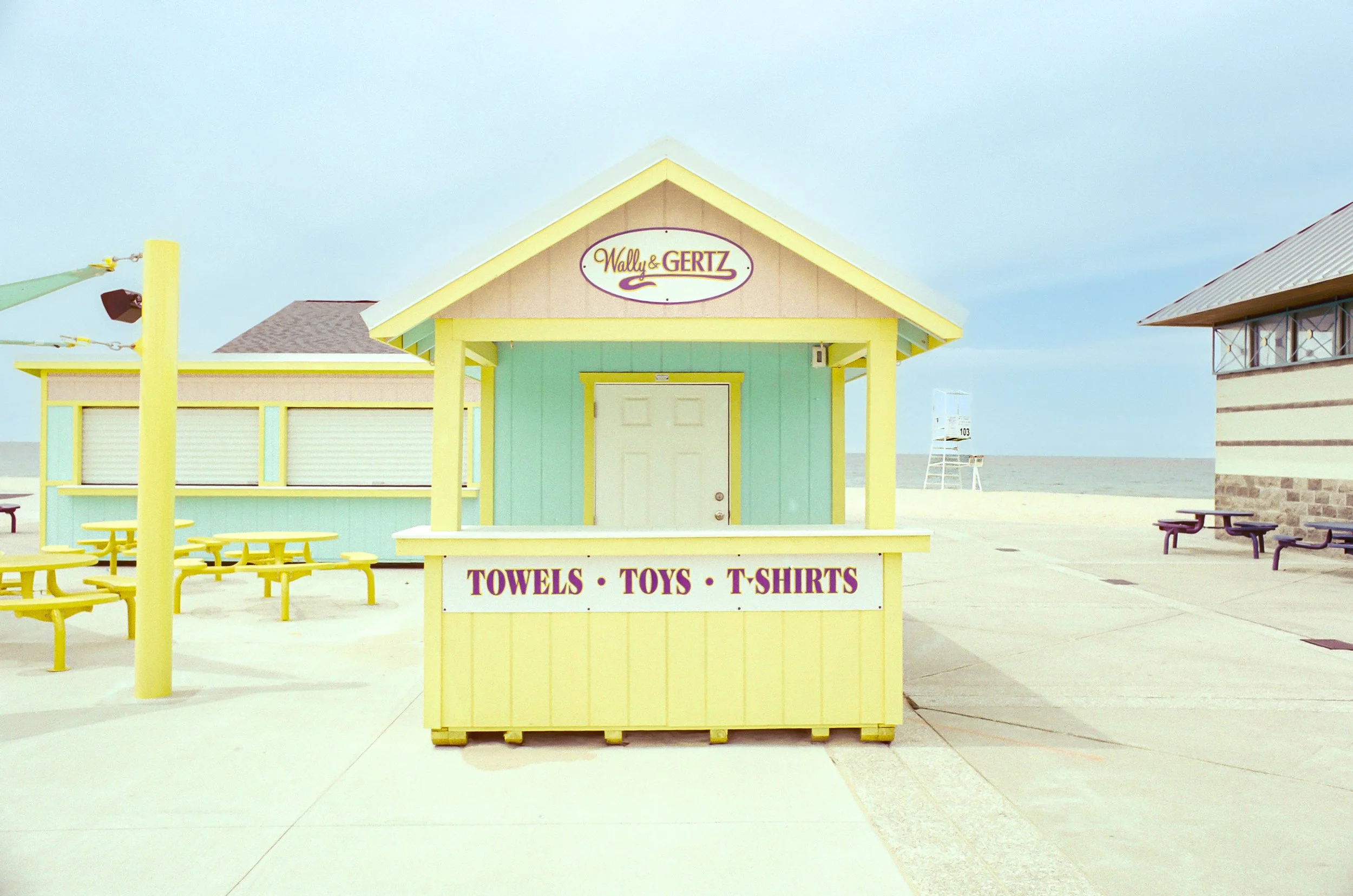 A small yellow stand on a beach with signs for towels, toys, and T-shirts, and a building behind it with pastel colors and a sign reading 'Wally & Gertz'.