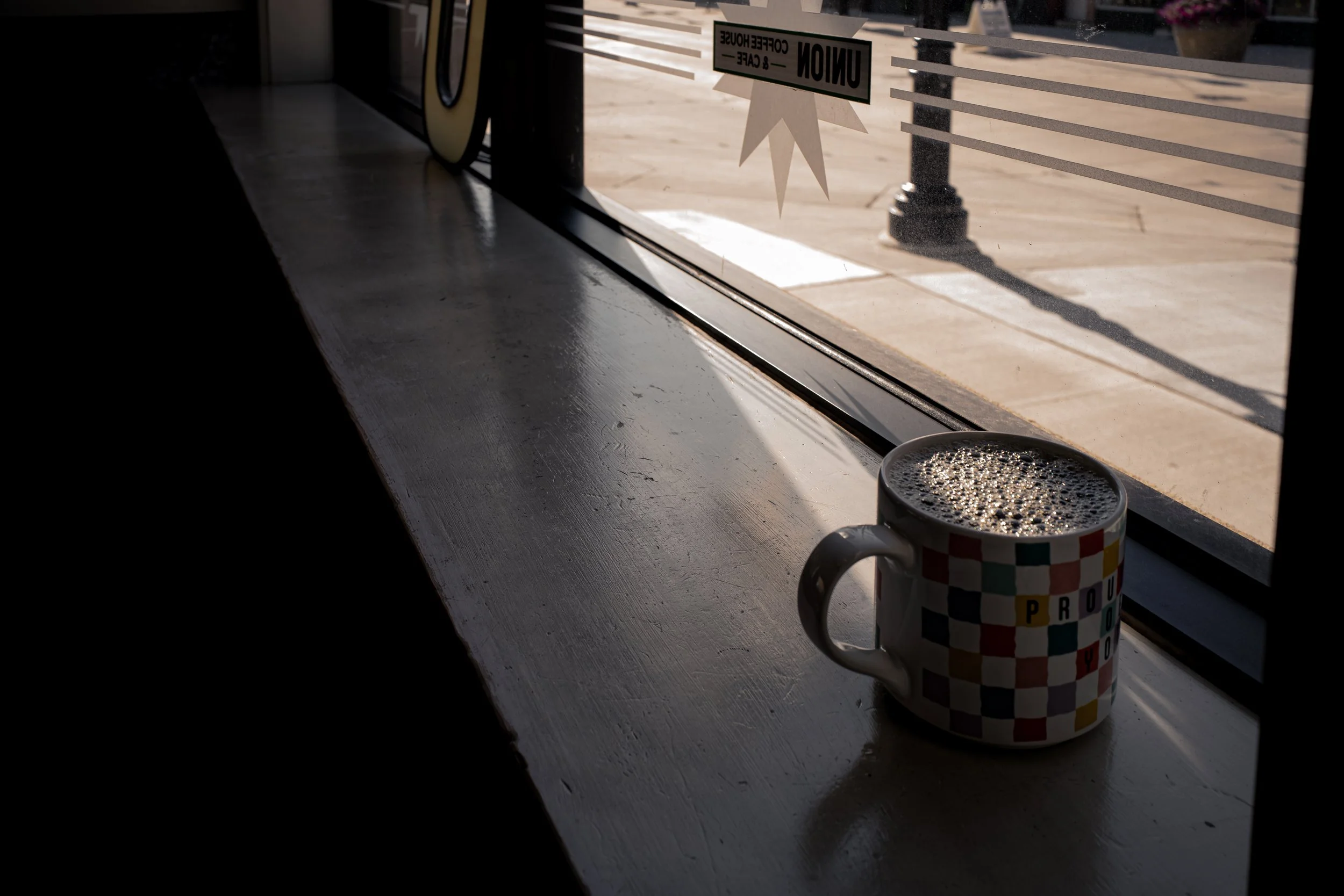 A checkered mug with a hot beverage on a windowsill, sunlight casting shadows outside with a 'Caution' sign on the window.
