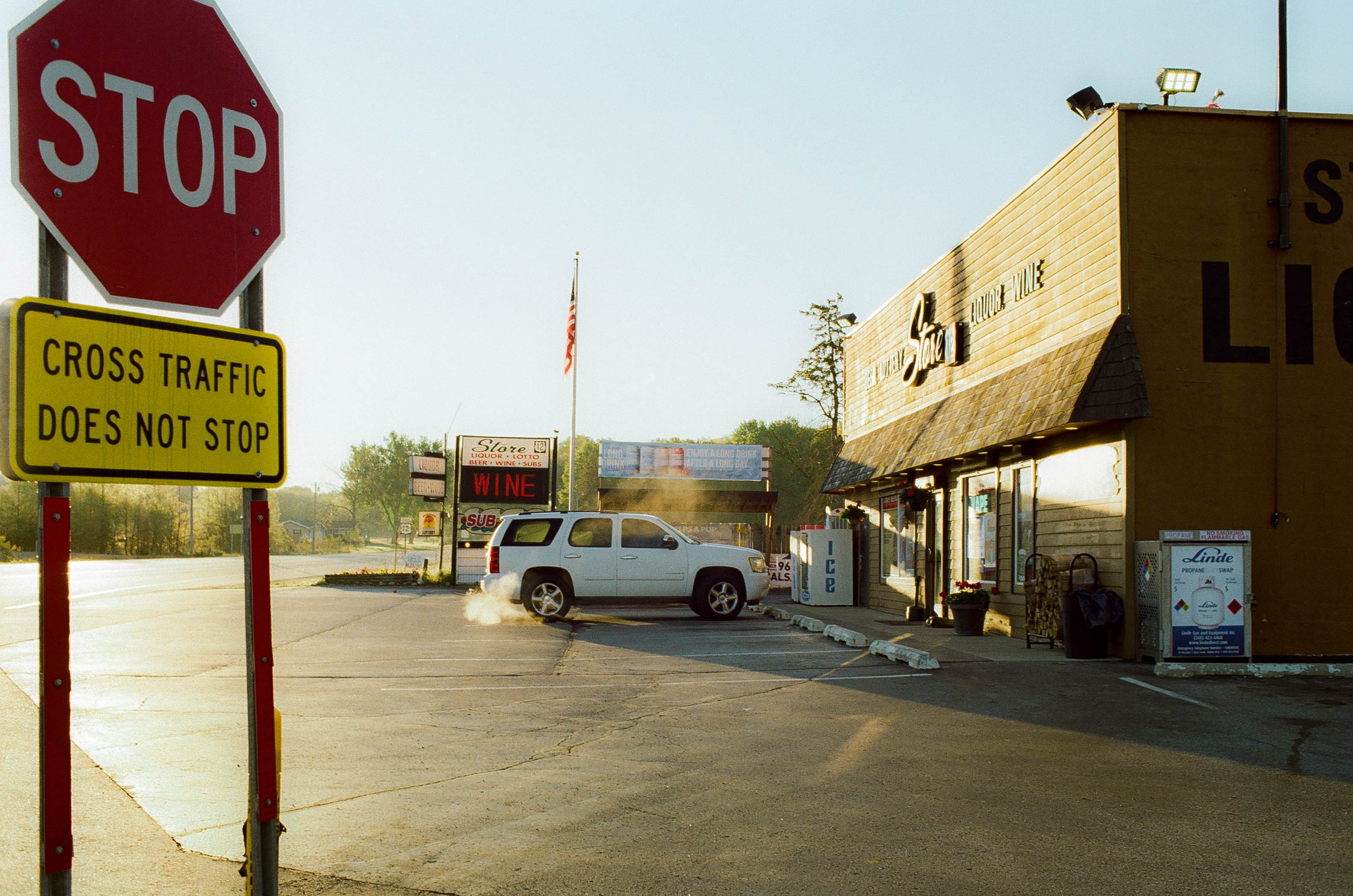 A parking lot outside a liquor store with a stop sign and a yellow cross traffic sign. A gray SUV is driving through, creating smoke. The store has a sign that reads "Store Liquor Lotto" and there are ice machines outside. The building is beige with 