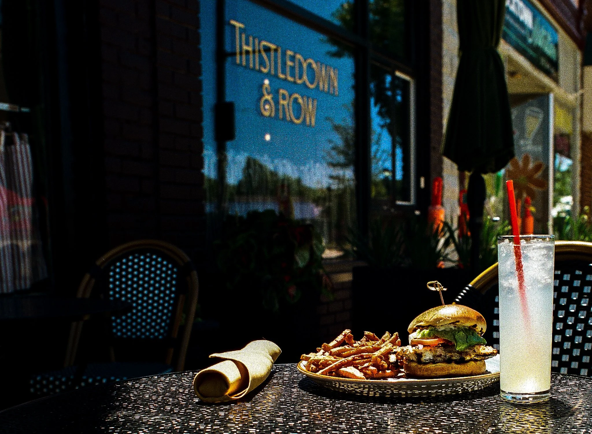A plate with a burger and fries on a black textured table outside at dusk, with chairs and windows from a restaurant in the background.