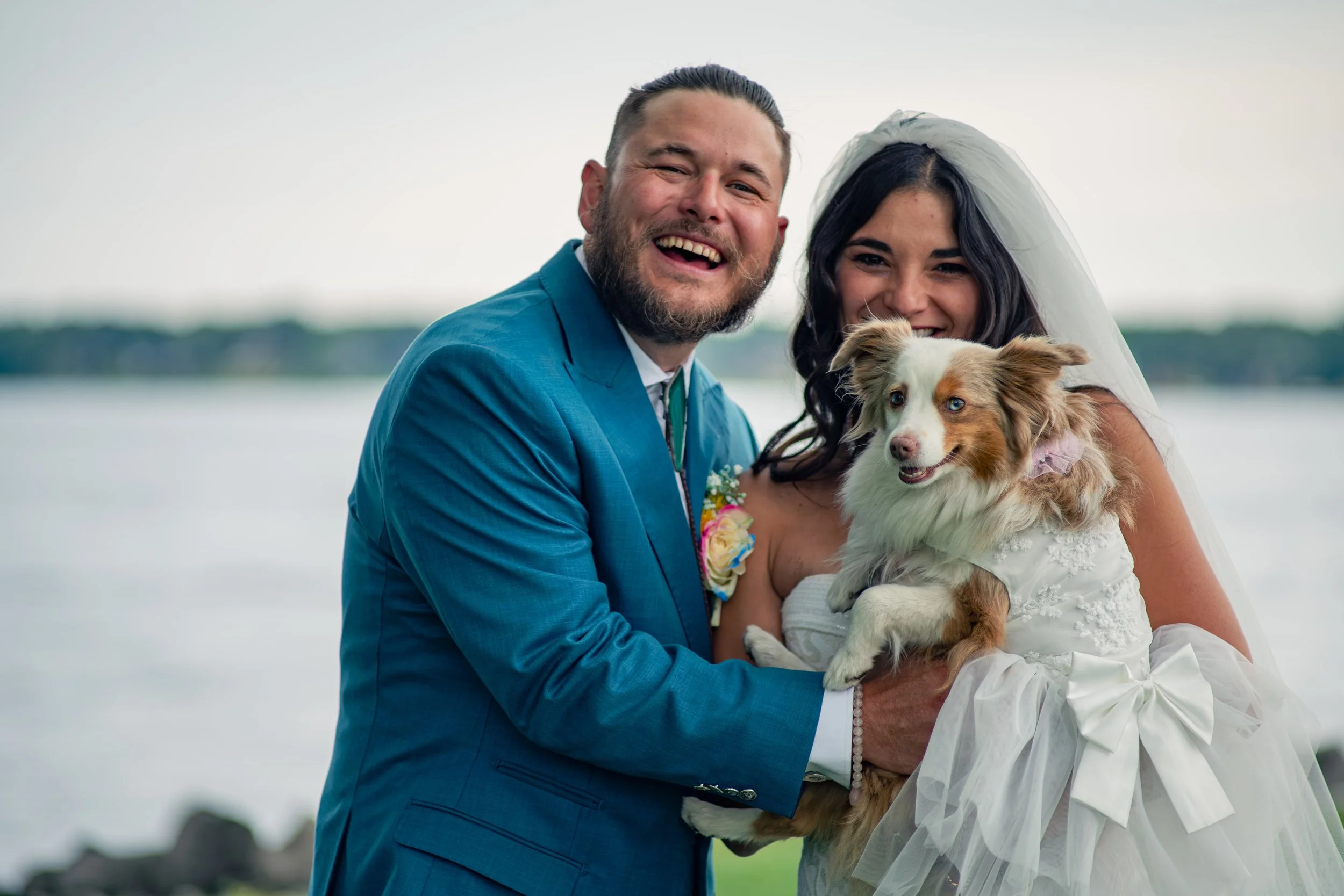A happy couple dressed in wedding attire holding a small dog outdoors near a body of water with a distant shoreline.