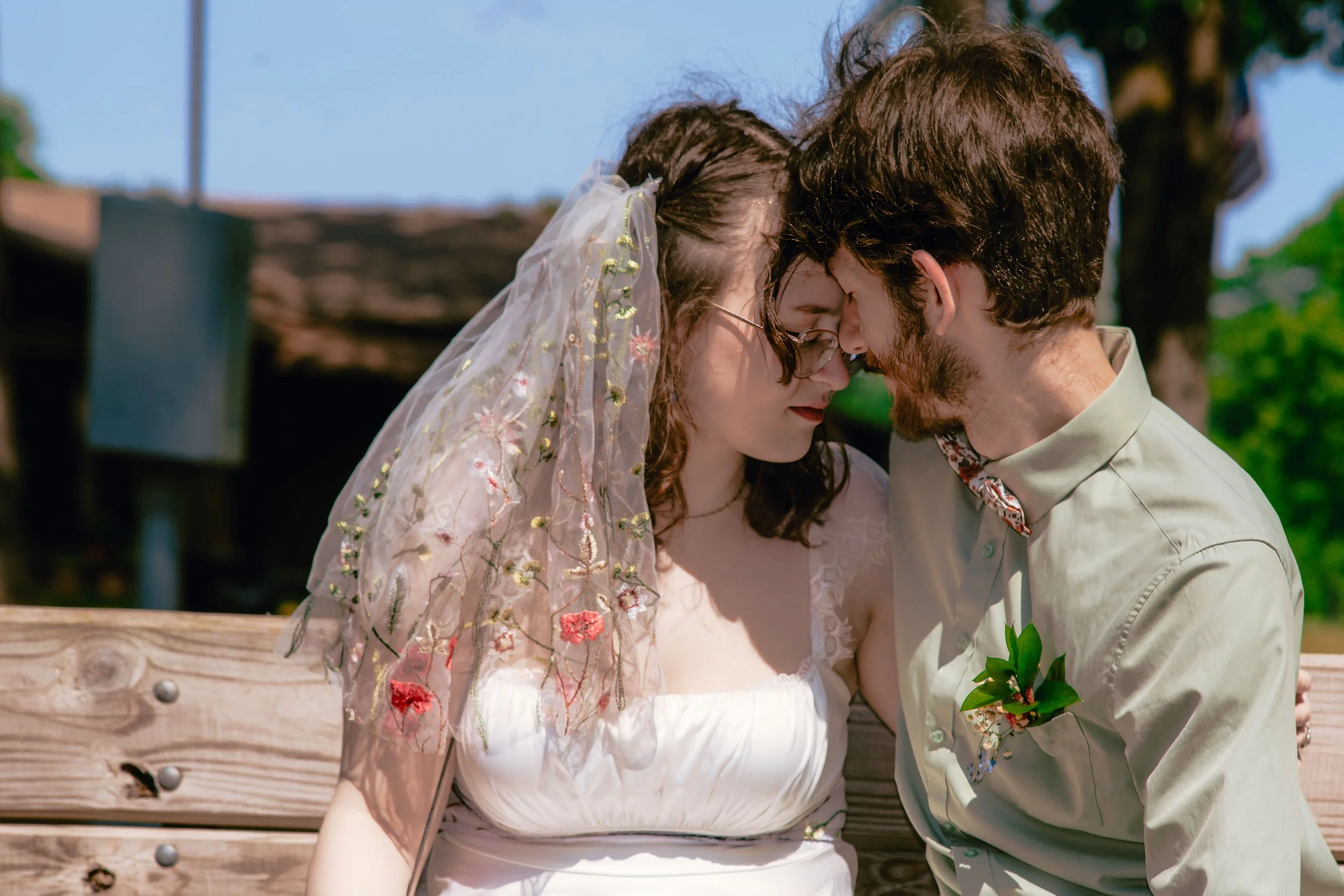 A man and woman are touching foreheads and holding hands, sitting on a wooden bench outdoors on a sunny day.