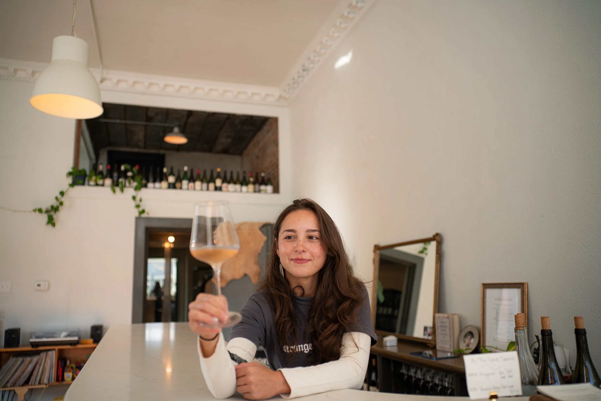 A woman with long brown hair and freckles sitting at a table in a restaurant, holding a glass of wine, smiling gently. The restaurant has a white wall with a row of wine bottles on a ledge in the background, some green plants, and warm lighting inclu