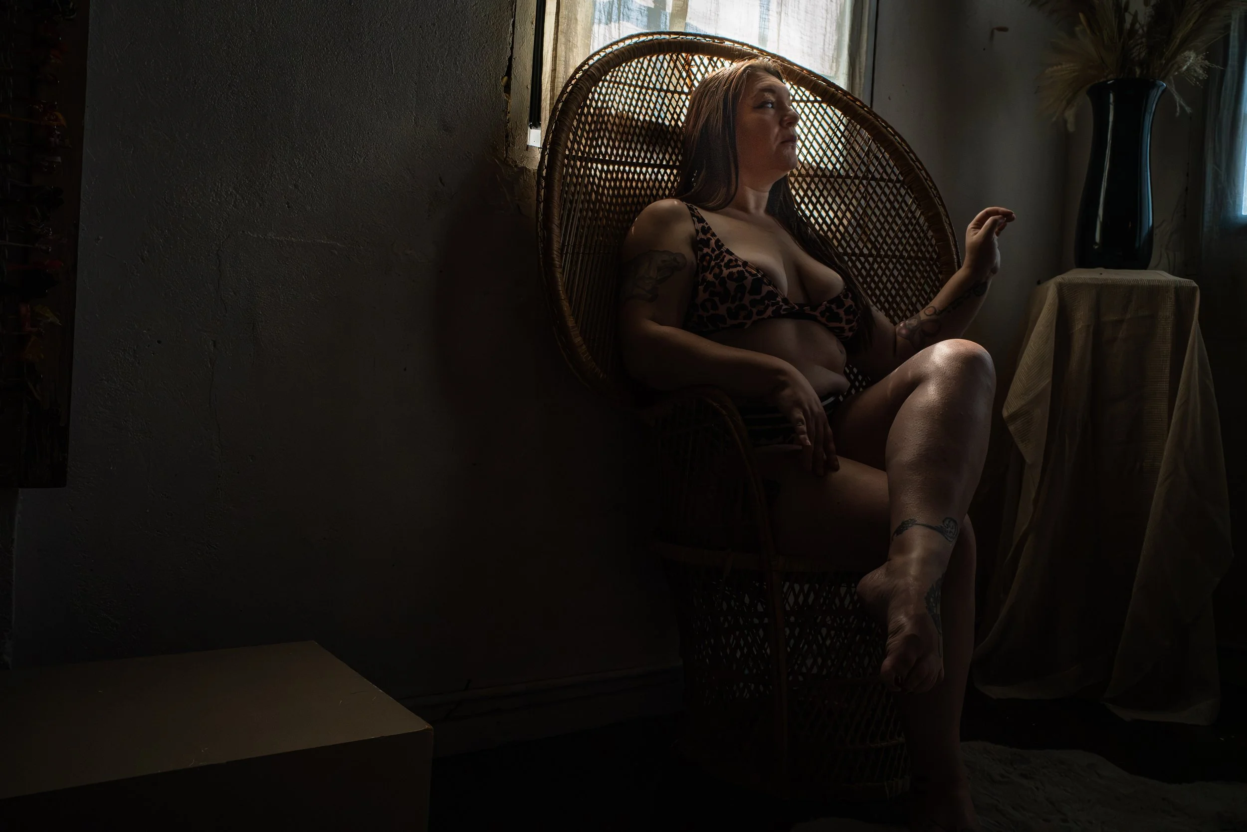 A woman sitting in a wicker chair, wearing a leopard print top, inside a dimly lit room with a small window behind her and a large vase with dried plants on a small table to her right.