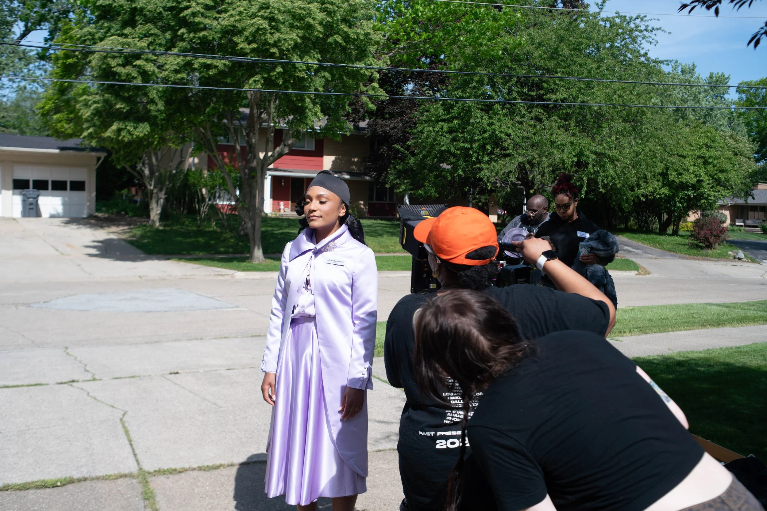 A young woman dressed as Dorothy from The Wizard of Oz standing outdoors during filming, surrounded by crew members and camera equipment, with trees and residential houses in the background.