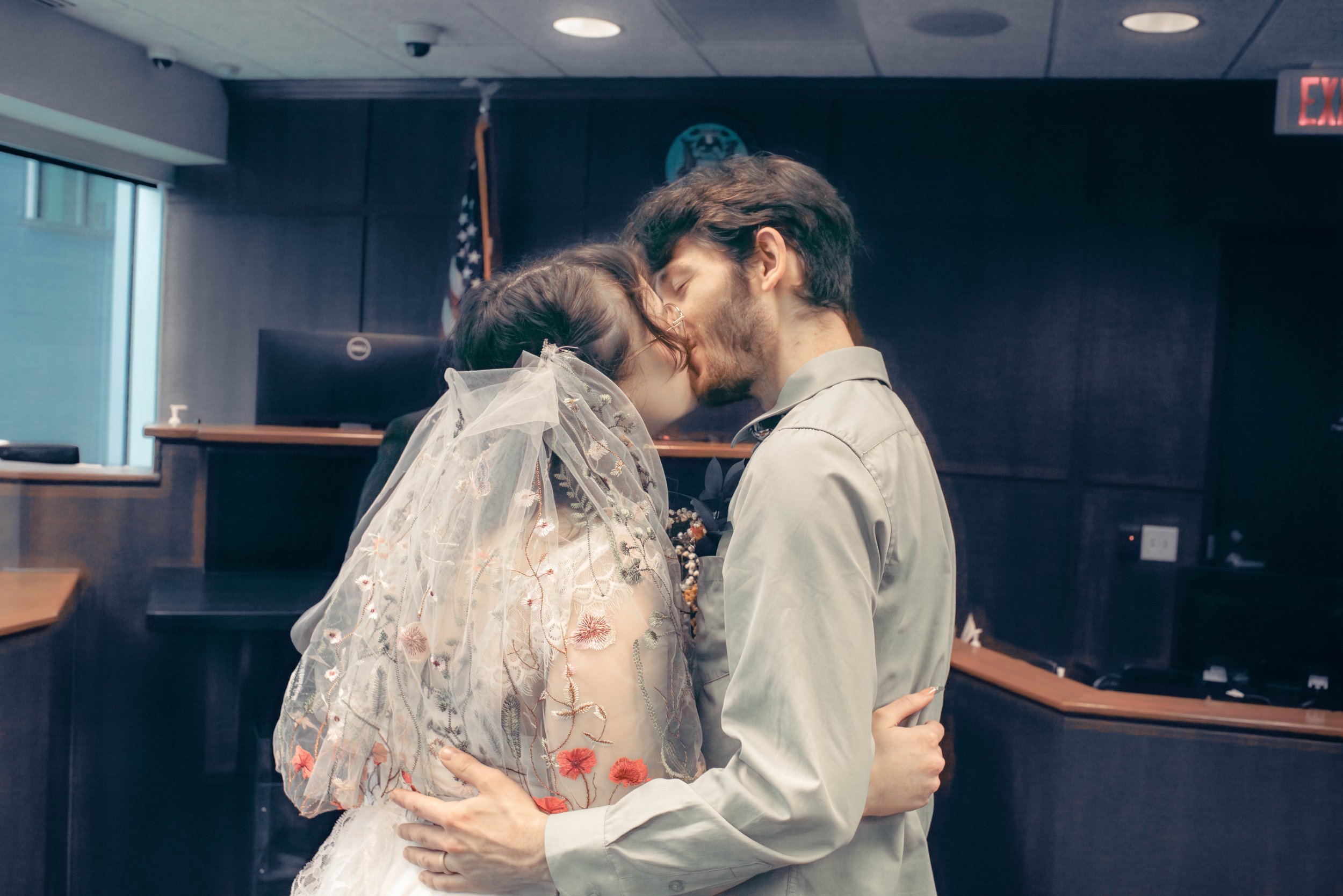 A couple is embracing and kissing each other in a room with dark walls, an American flag, and an emergency exit sign visible in the background.