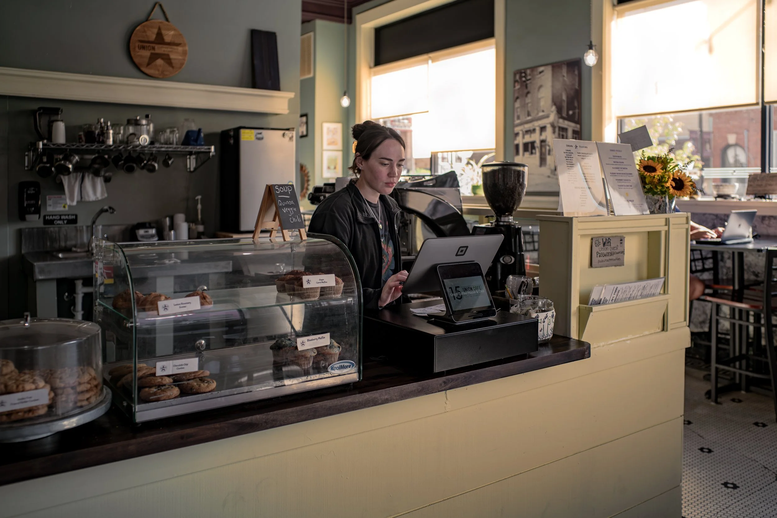 A woman working at a cafe counter with baked goods in glass display case, menu and flowers on the counter, and sunlight coming through large windows.