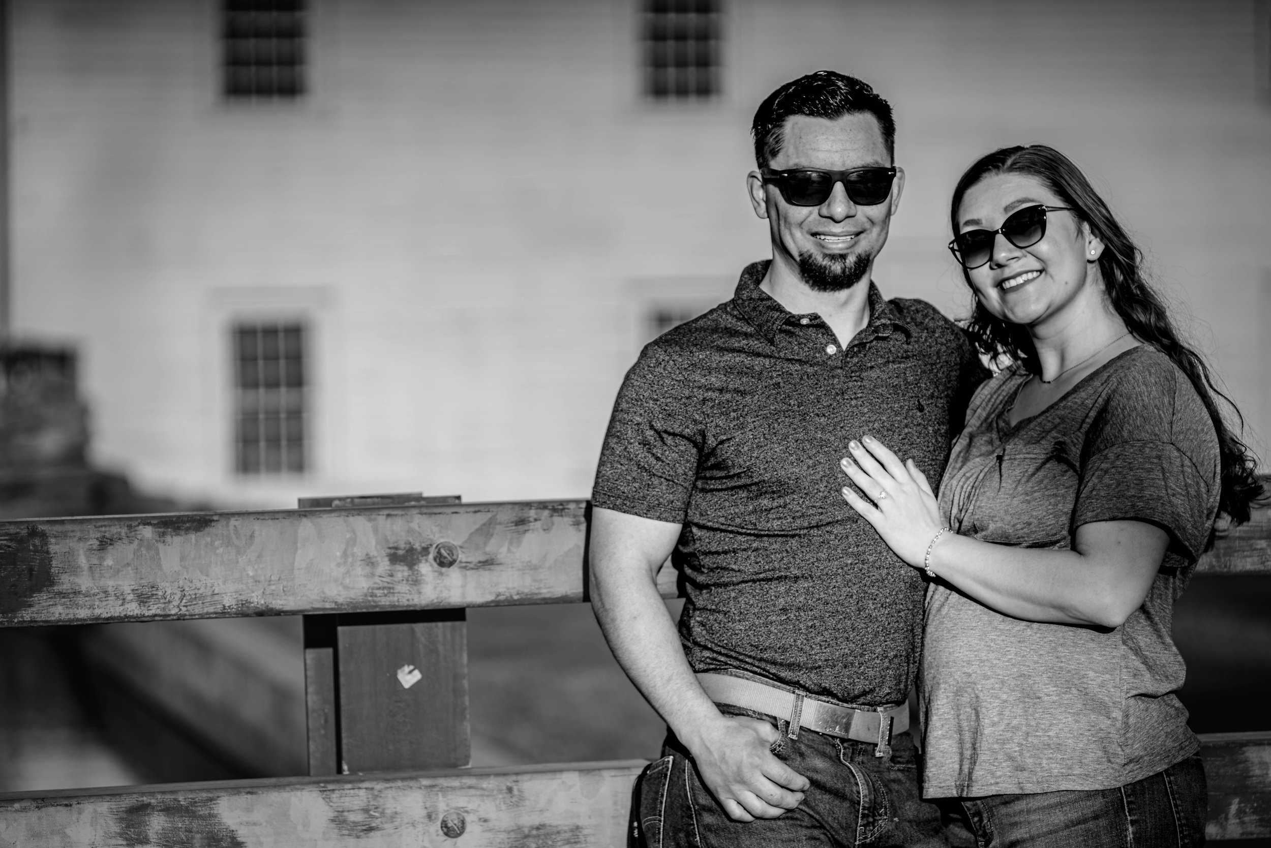 A smiling couple wearing sunglasses standing close together outdoors. The man has dark hair, a beard, and is wearing a patterned shirt. The woman has long hair, earrings, and is wearing a dark top. They are near a wooden railing with a building in th