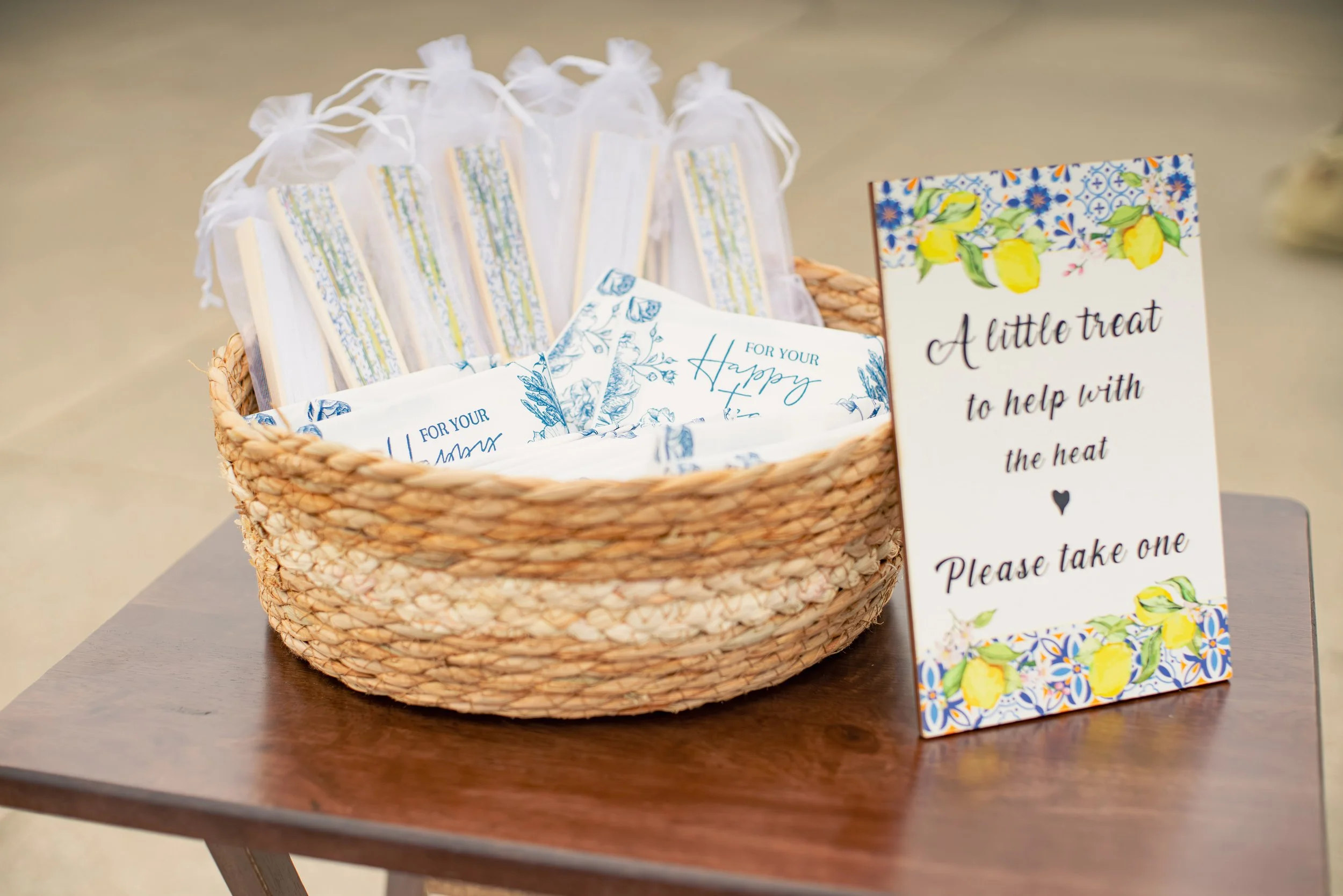 A wicker basket filled with individually wrapped treats on a dark wooden table. A decorative sign next to the basket reads, 'A little treat to help with the heat. Please take one' with floral and lemon designs.