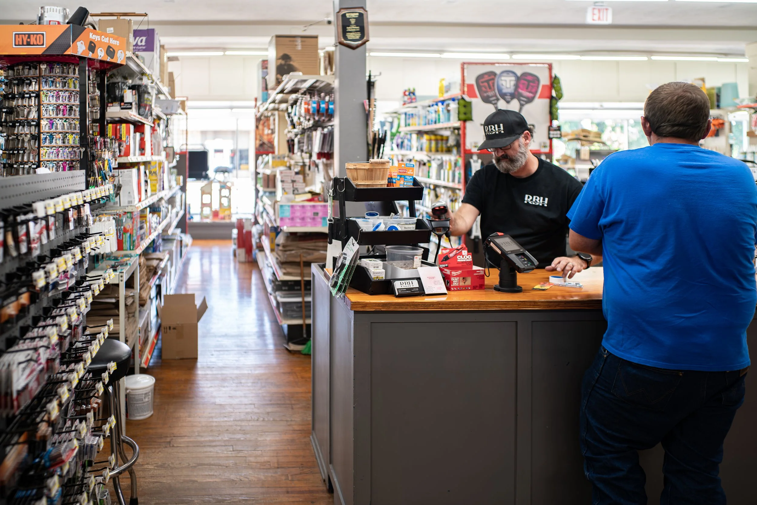 A store clerk at checkout counter interacting with a customer in a hardware store, with shelves of tools and supplies in the background.