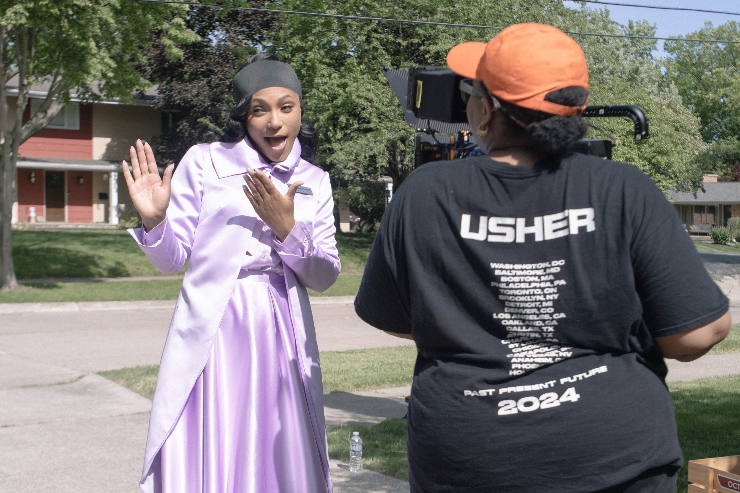 A woman in a purple dress and black headscarf is talking to a camera operator wearing a black T-shirt with 'USHER' on the back and an orange cap. The woman appears surprised or excited, with her hand near her face, and is outdoors with trees and hous