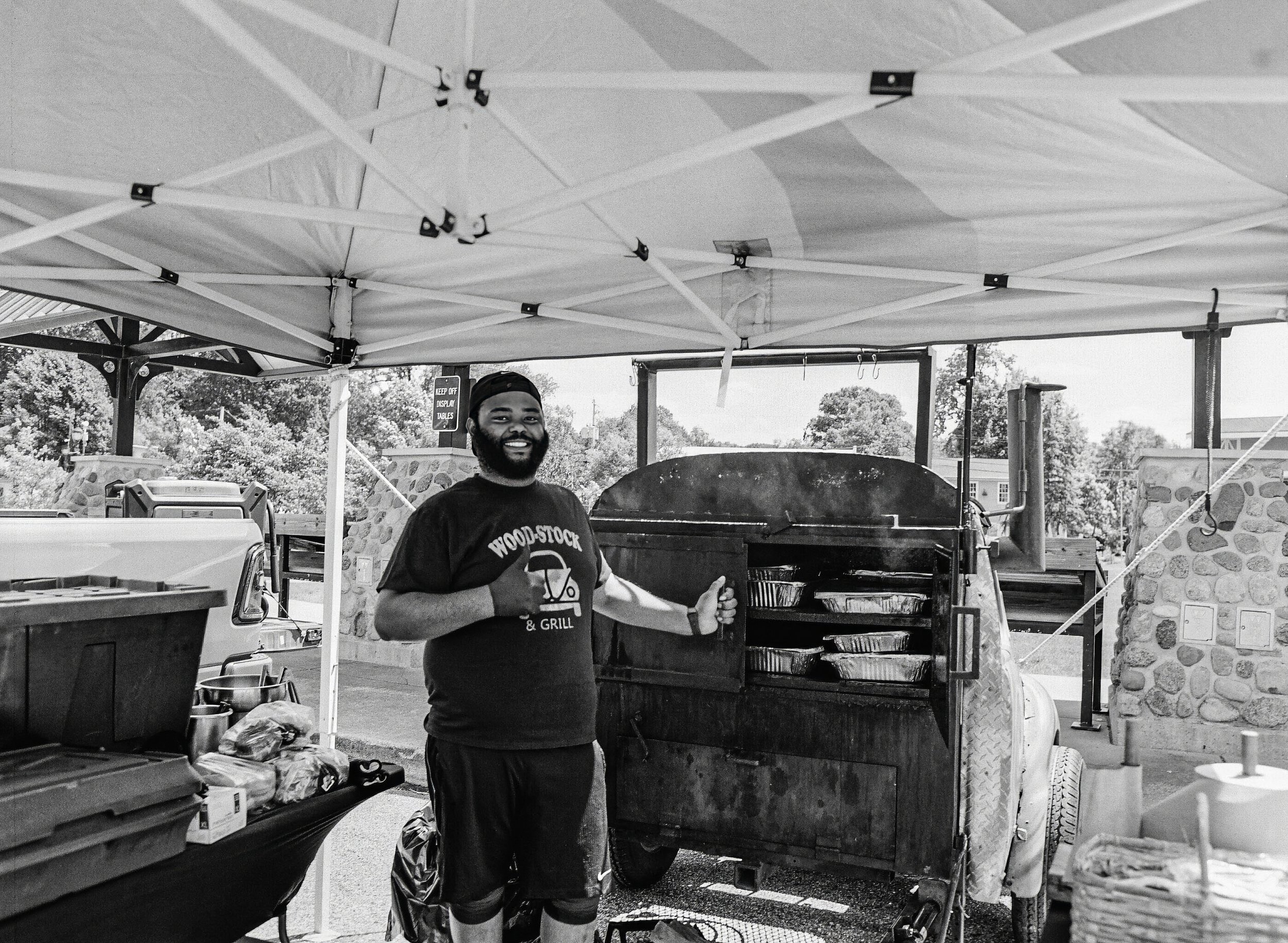 A man with a beard and a cap is standing behind a barbecue grill at an outdoor event, smiling and giving a thumbs-up. There is a canopy tent overhead and various cooking and outdoor equipment visible around him.