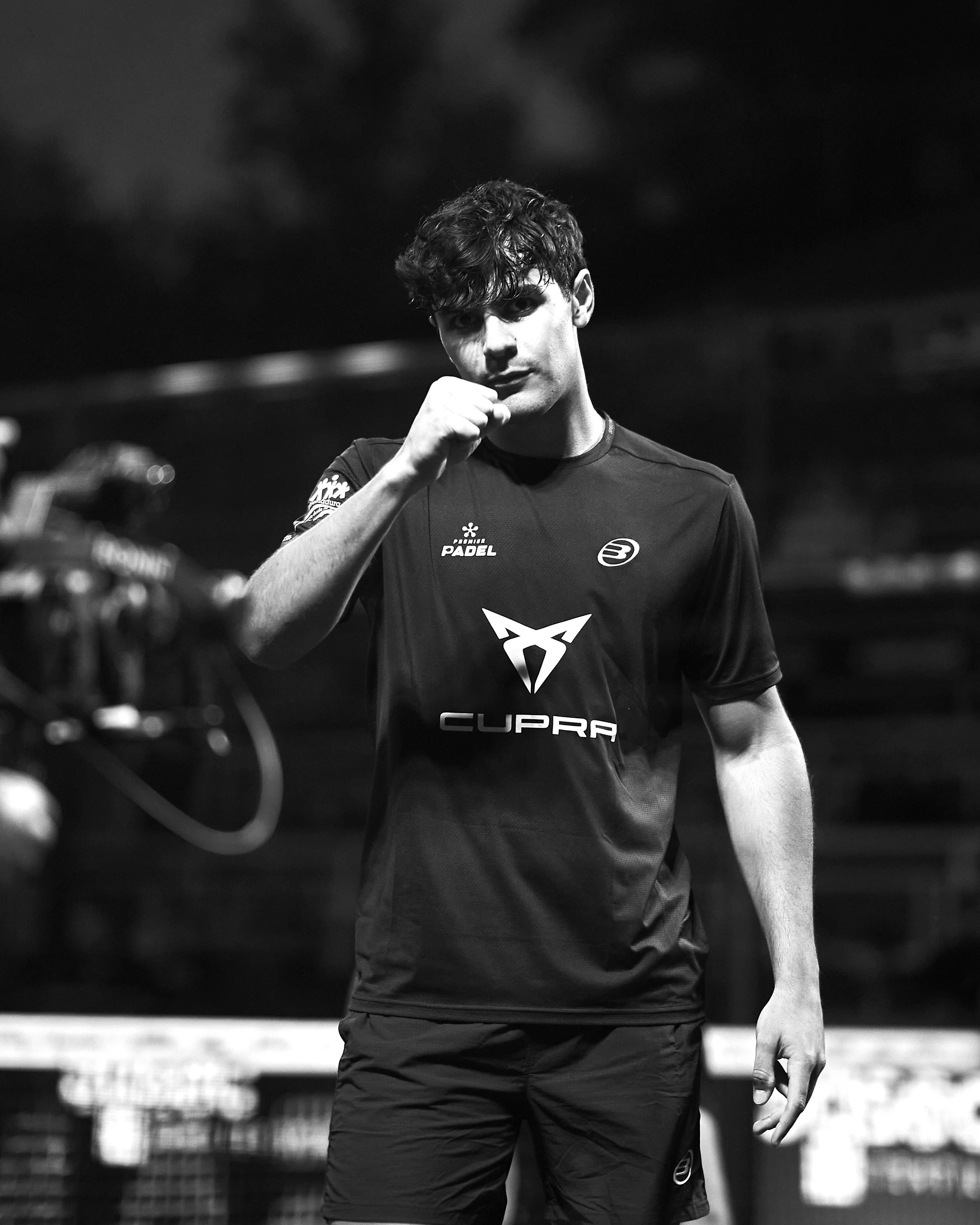 Black and white photo of a young man in sports attire with a serious expression, fist near his chin, standing outdoors at night.