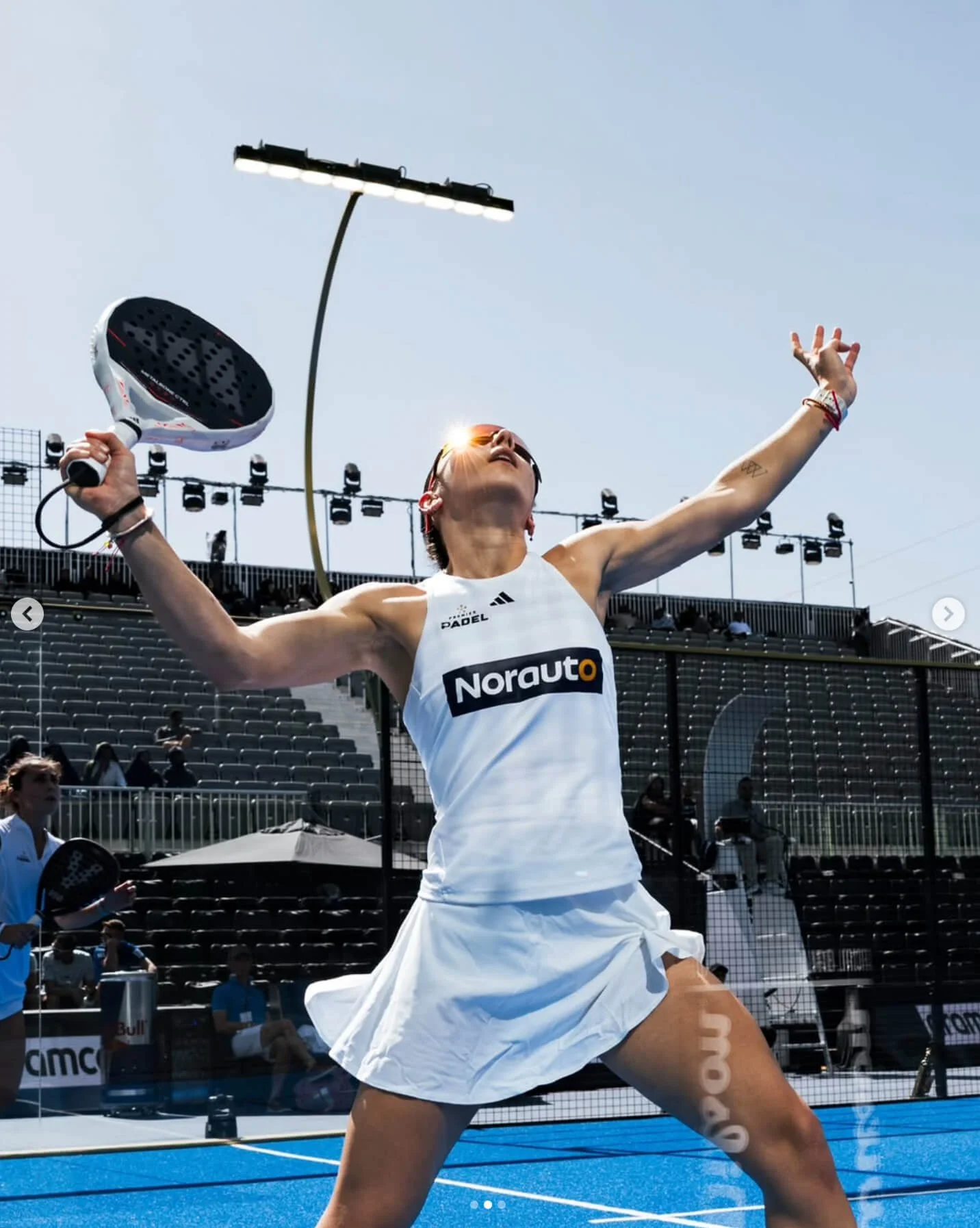 A female padel player in a white sports outfit celebrating on a blue padel court, holding her racket aloft with one arm raised, in front of an empty stadium with some other players and officials in the background.