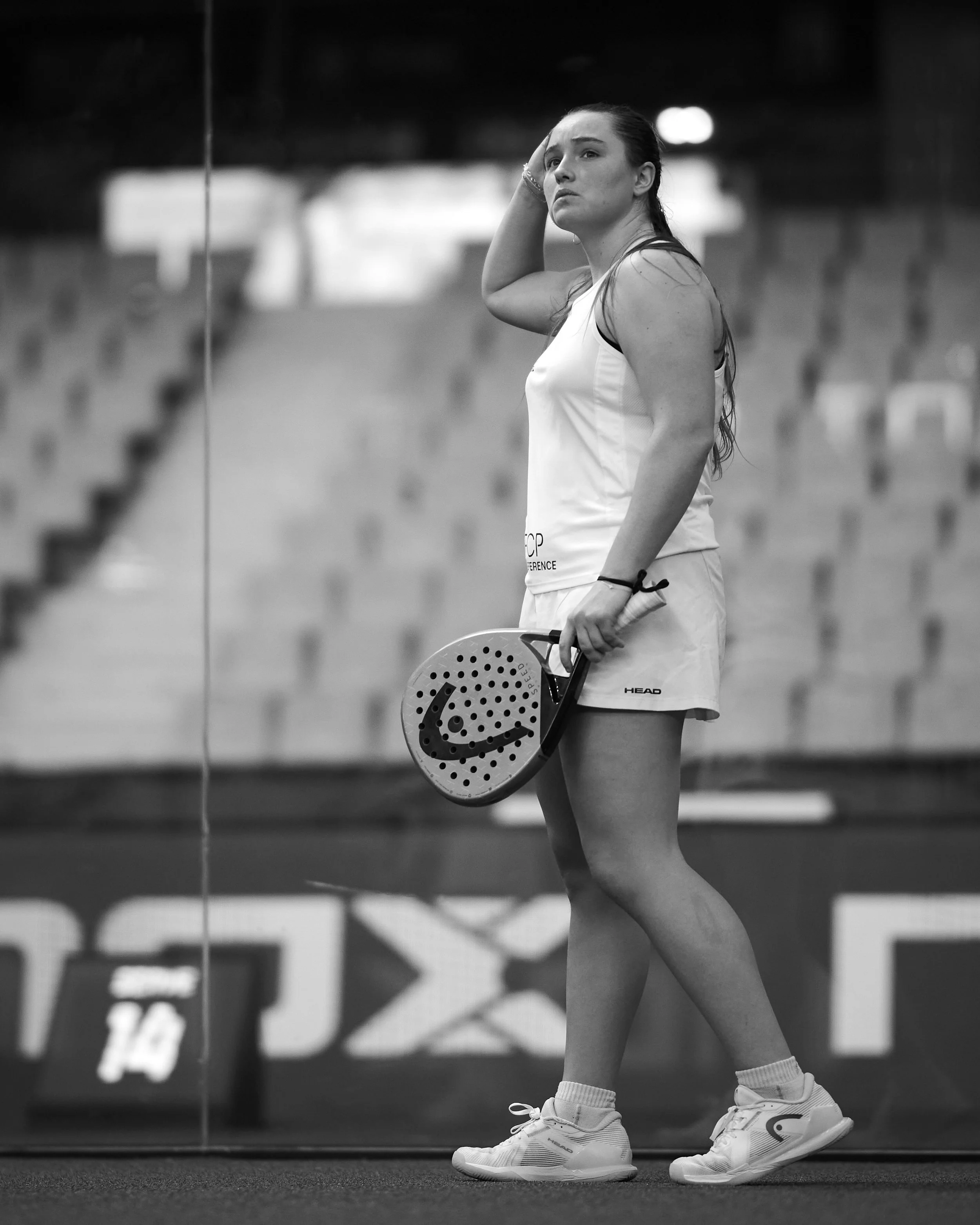 A female padel player standing on the court, holding a padel racket in her right hand, with her left hand on her head, wearing athletic shorts, a tank top, and tennis shoes, in an indoor padel facility.
